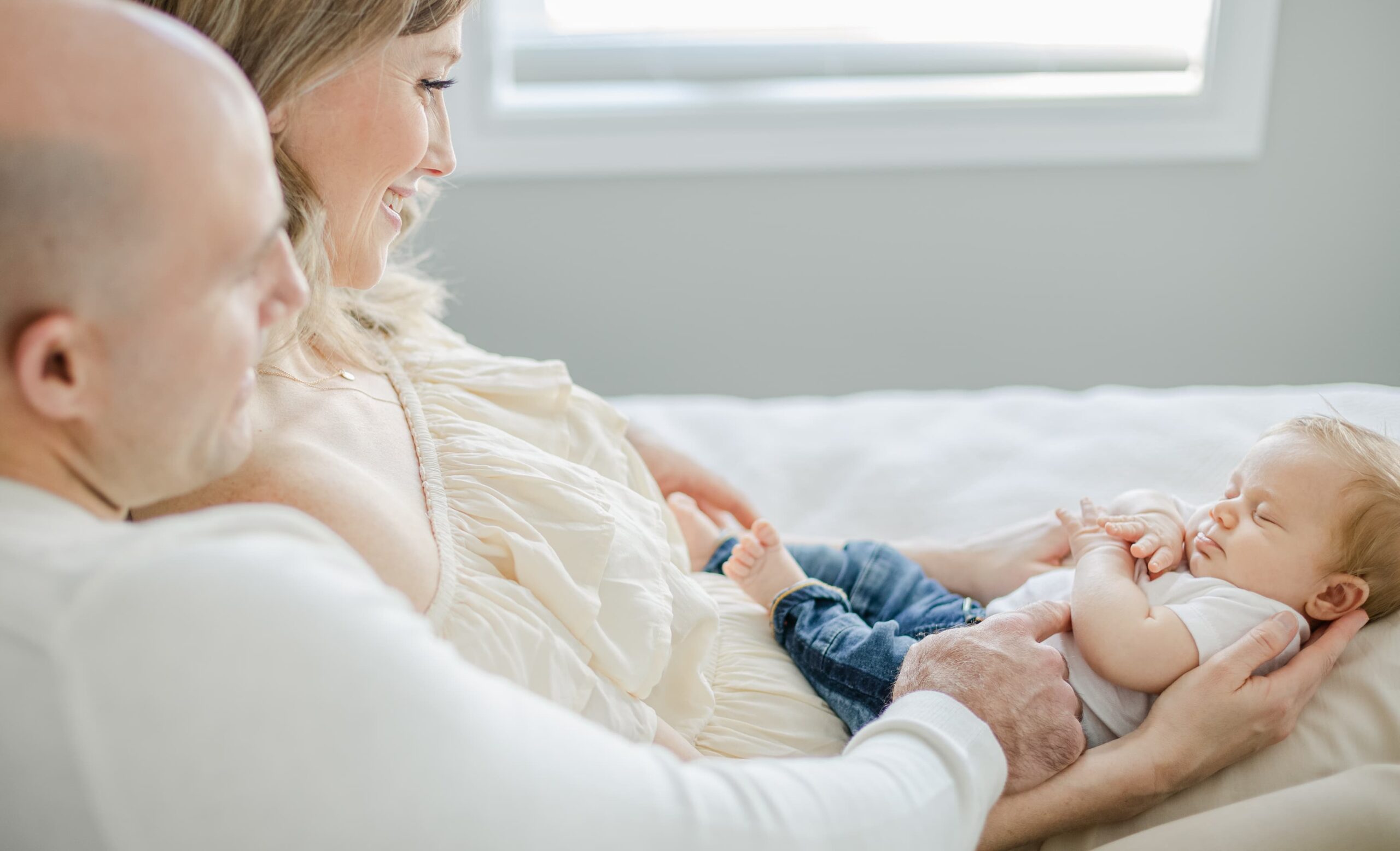 Happy parents sit on a bed with their sleeping newborn baby in mom's lap after visiting a baby boutique costa mesa