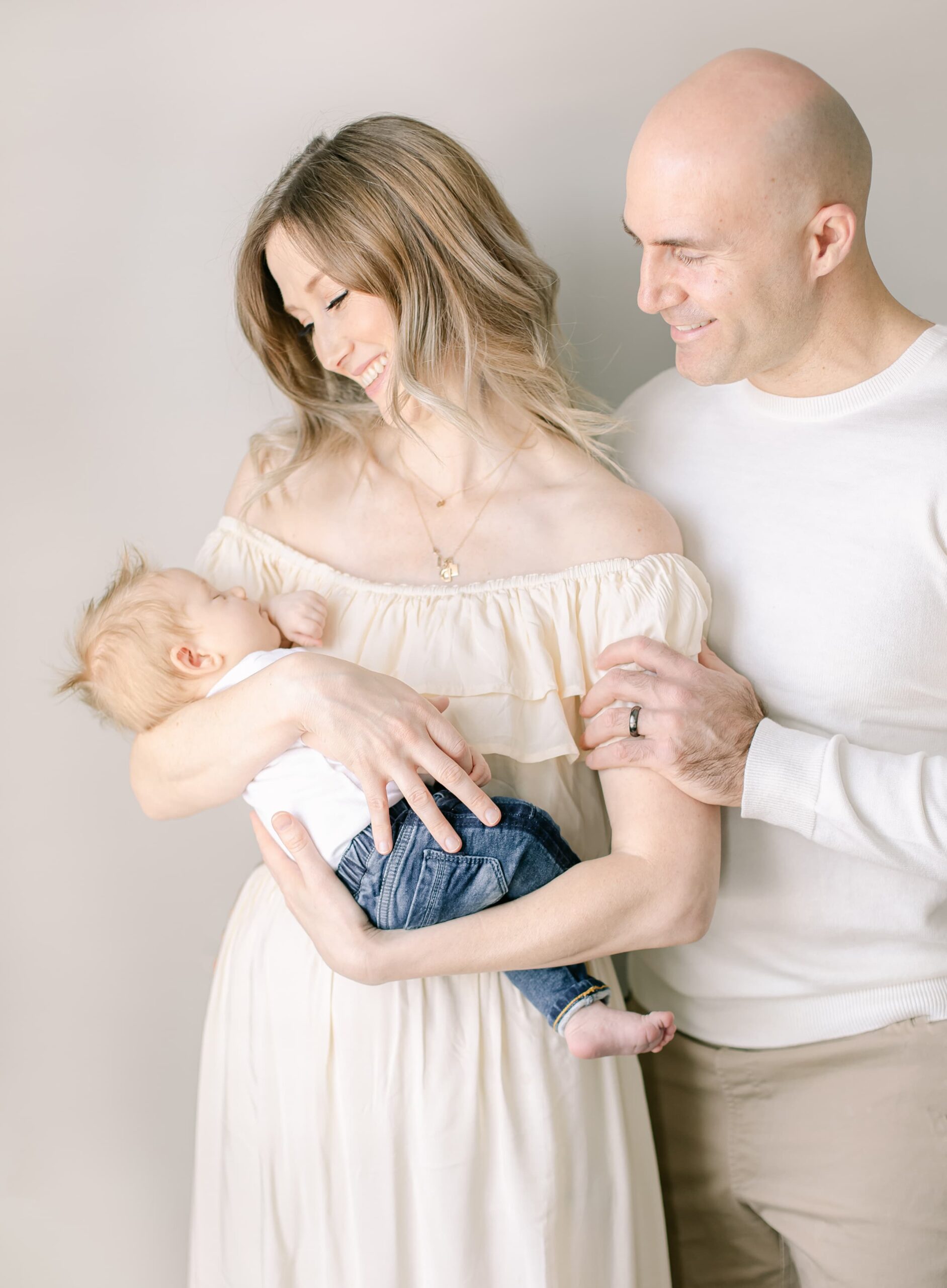 Happy new parents smile down at their sleeping newborn baby in mom's arms while standing by a window