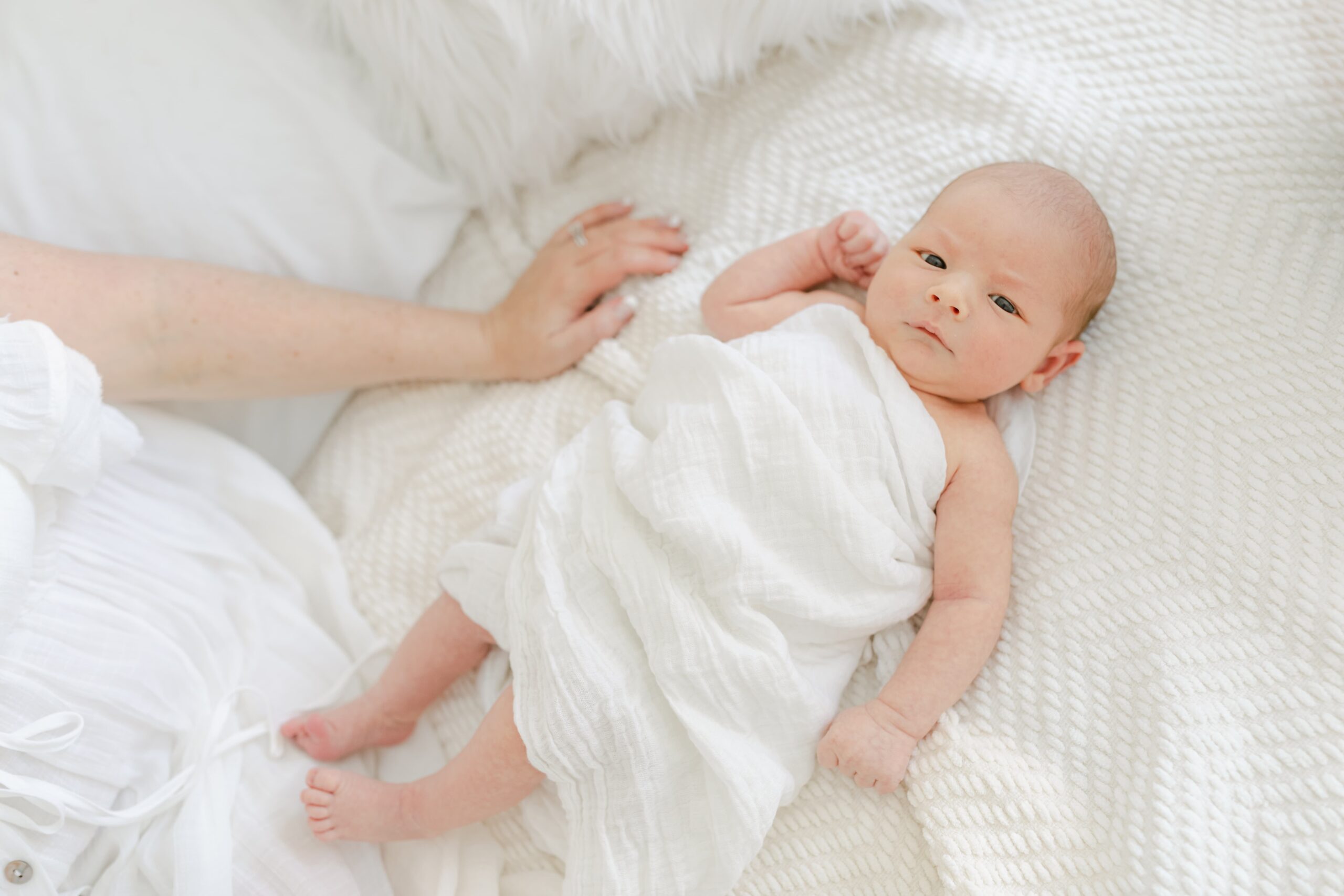 A newborn baby lays on a white bed wrapped in a white cloth with eyes open