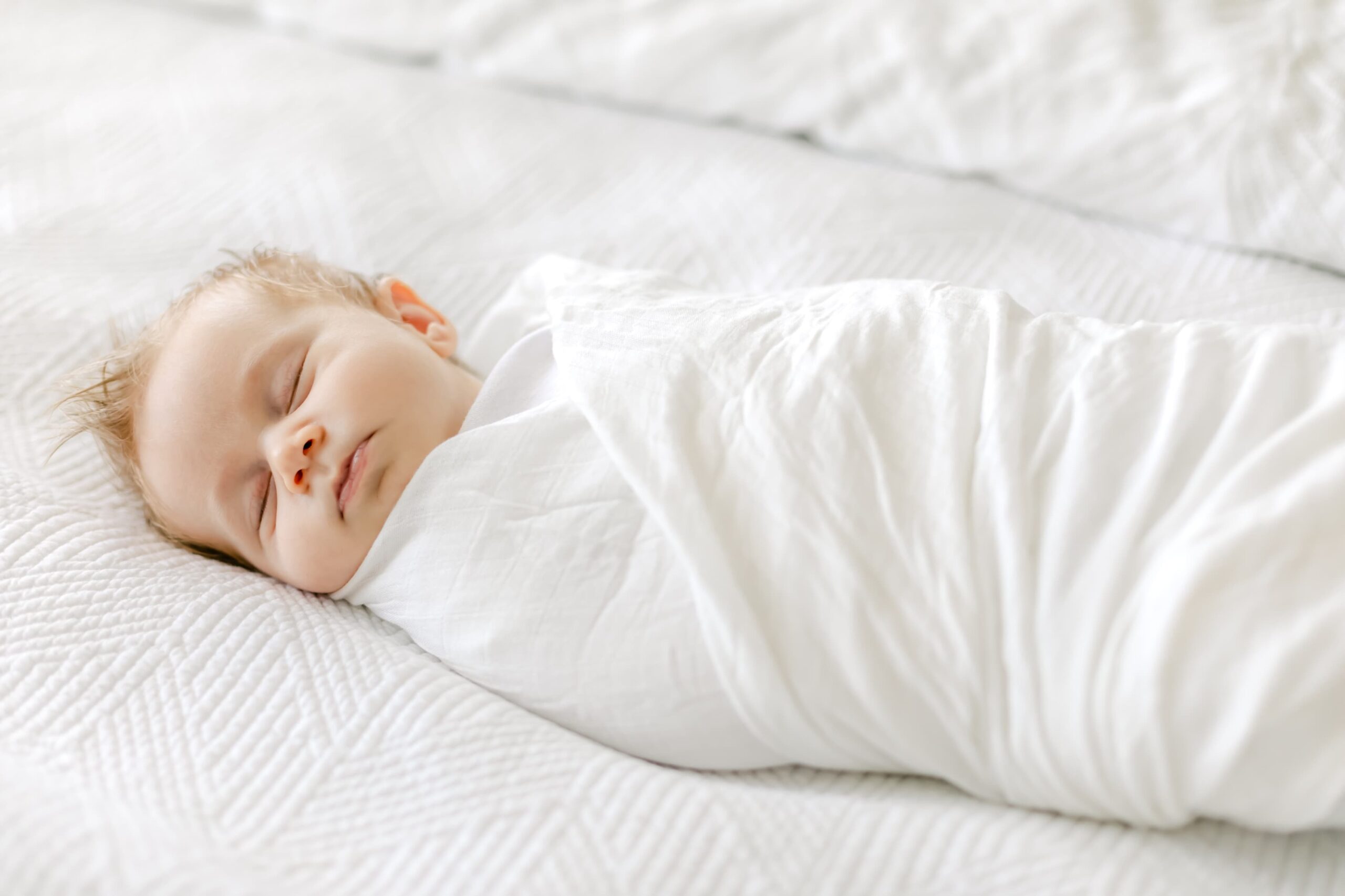 A newborn baby sleeps in a white swaddle on a bed before visiting a baby boutique costa mesa