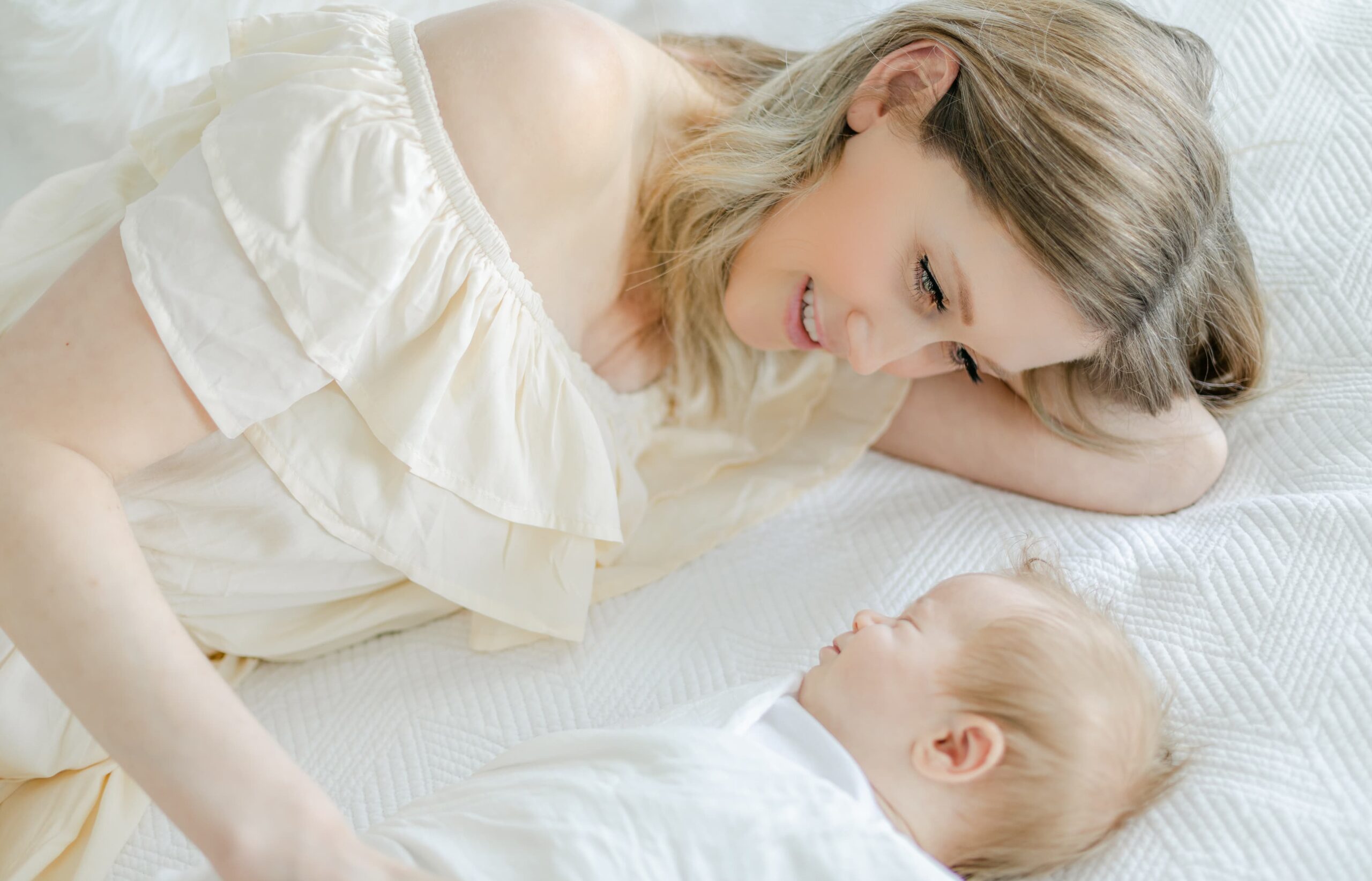 A mother in a cream dress lays on a bed smiling with her sleeping newborn baby after visiting a baby boutique costa mesa