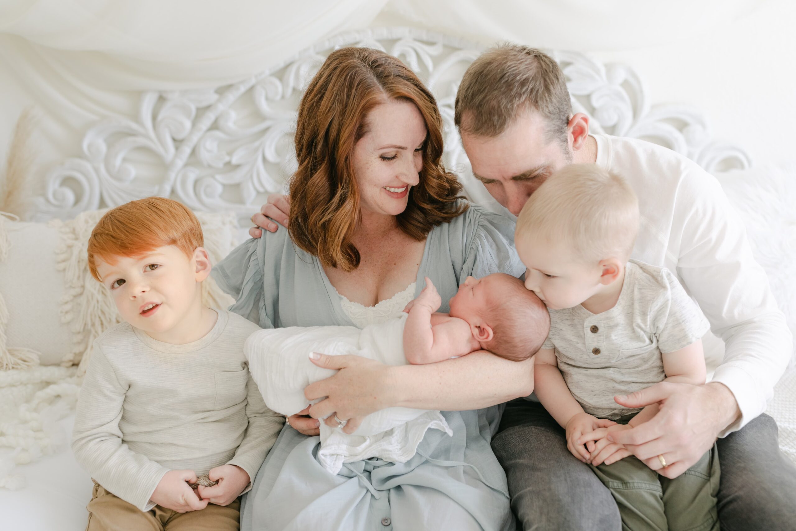 A toddler boy sits in dad's lap while kissing his newborn baby sibling being held by mom while his other brother sits at mom's side