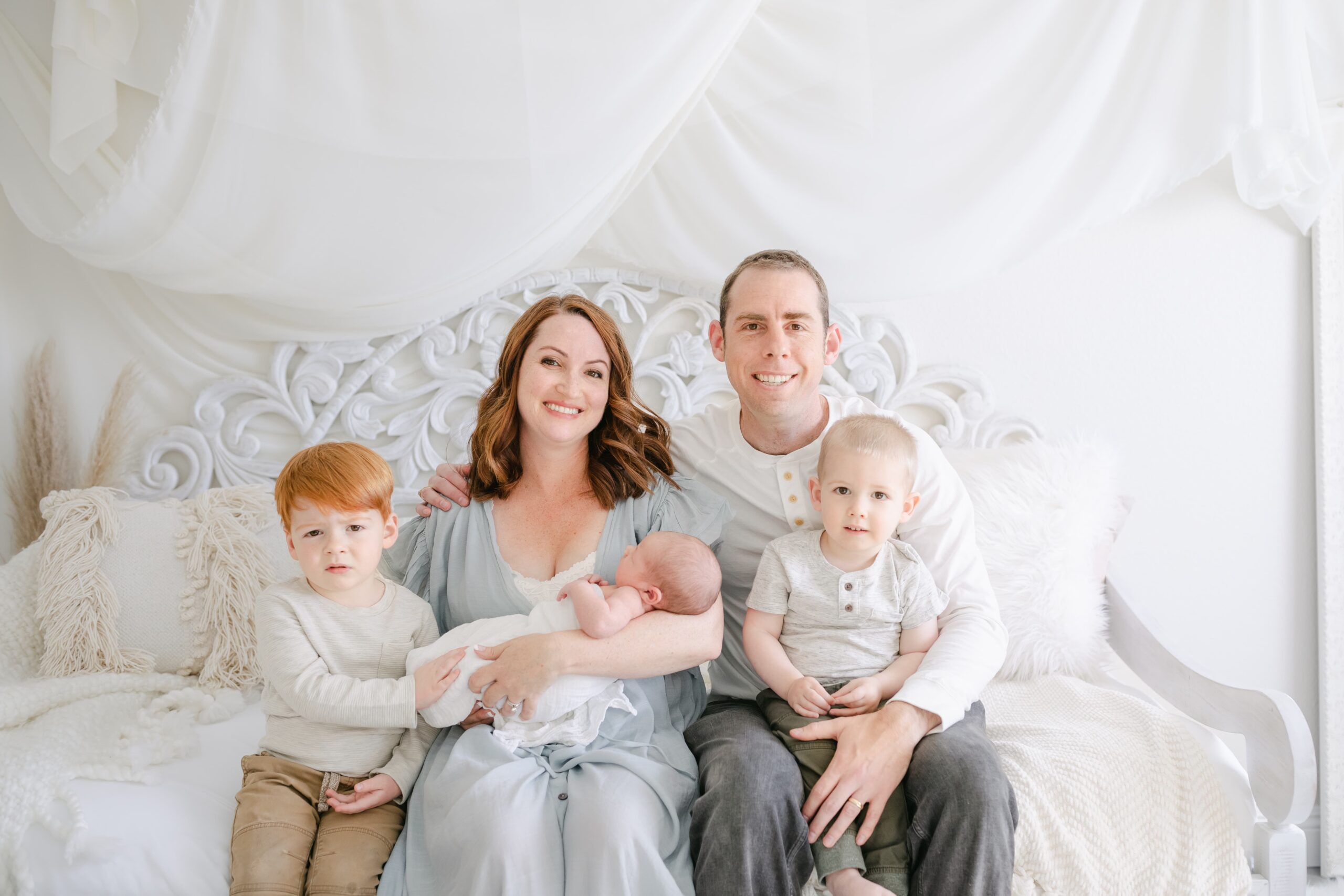 A mom and dad smile while sitting on a day bed in a studio with their newborn baby and two toddler sons