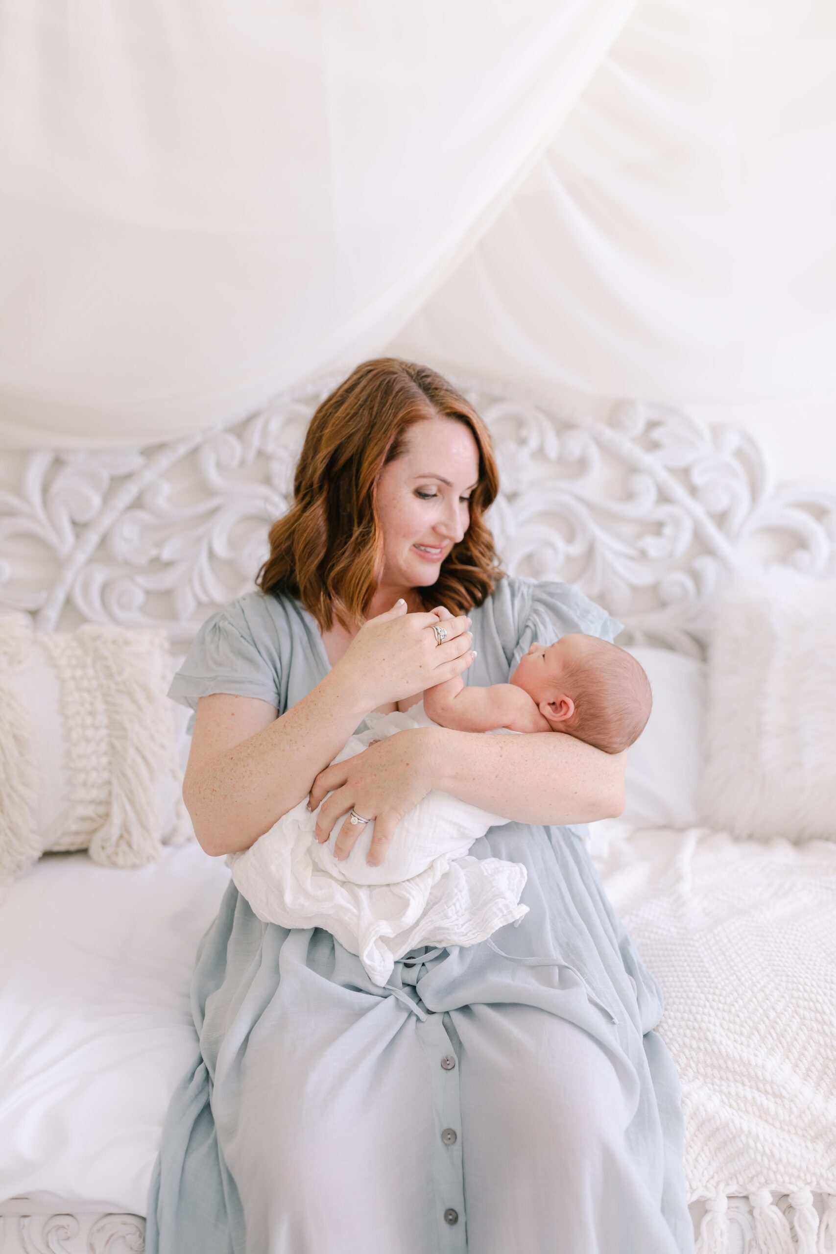 A happy mom sits on a daybed playing with her newborn baby before visiting baby boutique newport beach