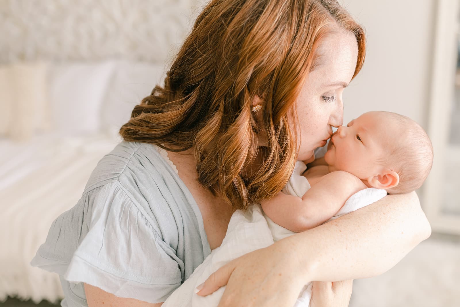 A mother in a blue dress kisses her newborn baby while standing in a bedroom after visiting a Baby Boutique Newport Beach