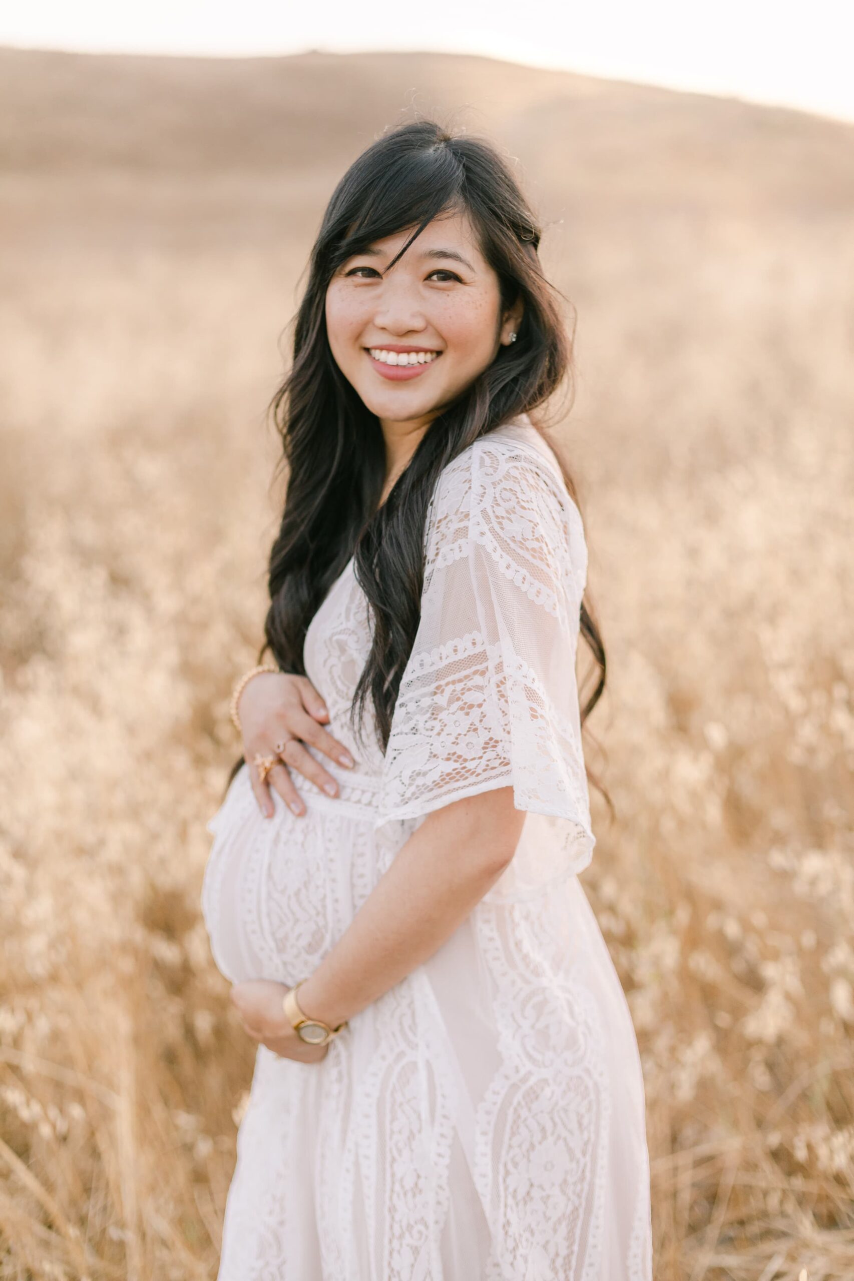 A mom to be walks through hills of golden grass in a white lace maternity dress