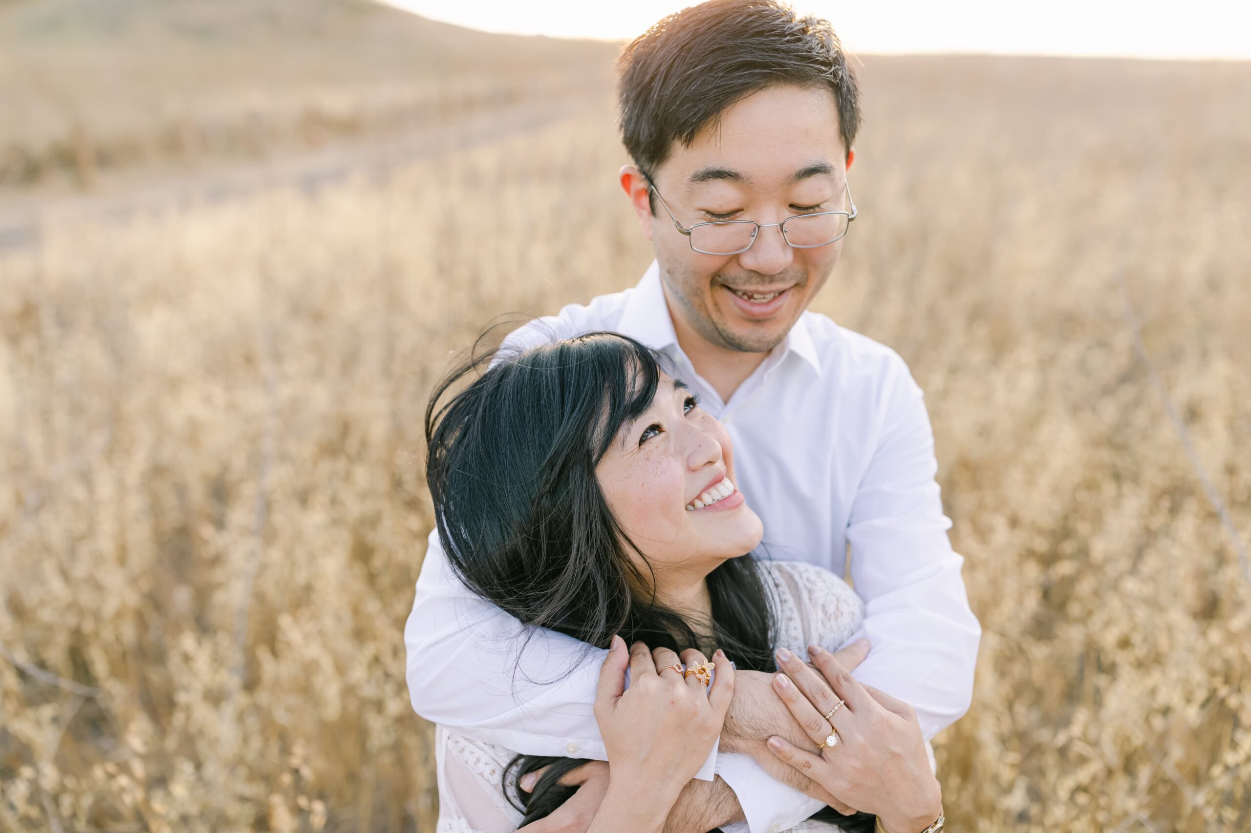 A mom to be leans into the chest and laughs with her husband in a field of golden grass after visiting baby shower venues orange county