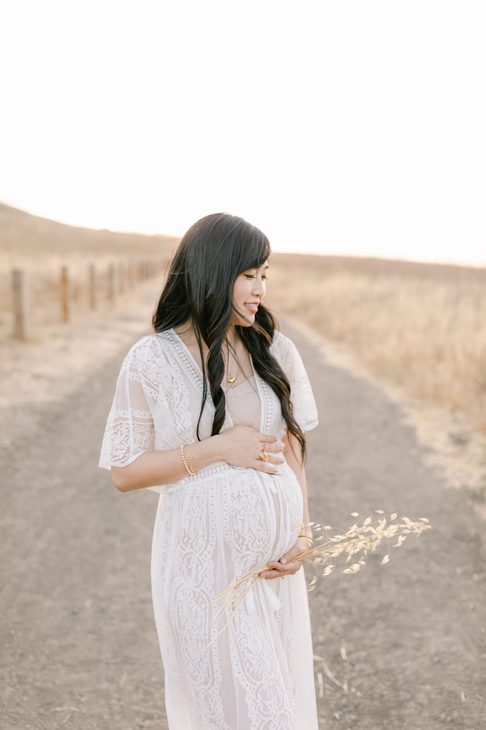 A mom to be in a white lace dress walks down a rural path through a field of tall golden grass after searching for baby shower venues orange county