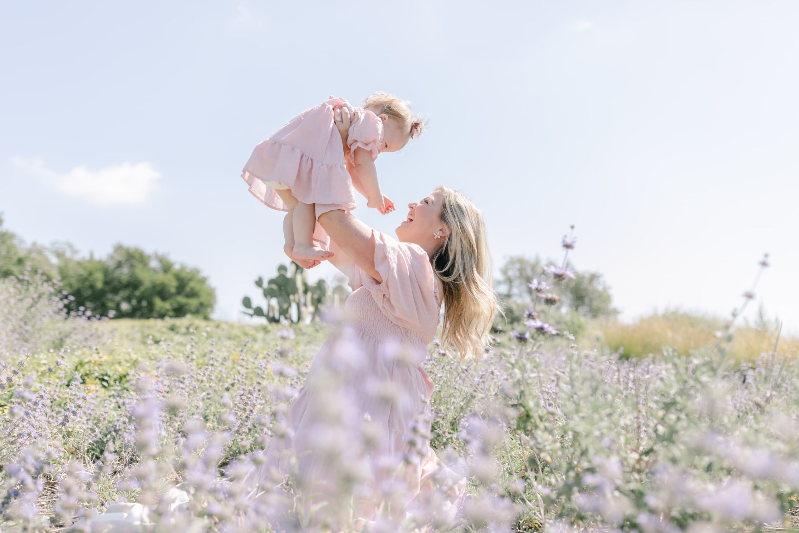 A mother in a pink dress lifts and laughs with her toddler daughter in a field of purple wildflowers after meeting with huntington beach pediatricians