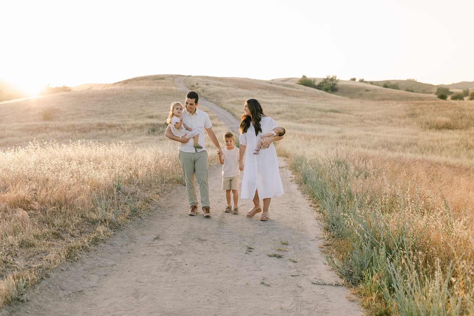 A mom and dad carry their newborn baby and toddler daughter while walking and holding hands with their toddler son in a field trail at sunset