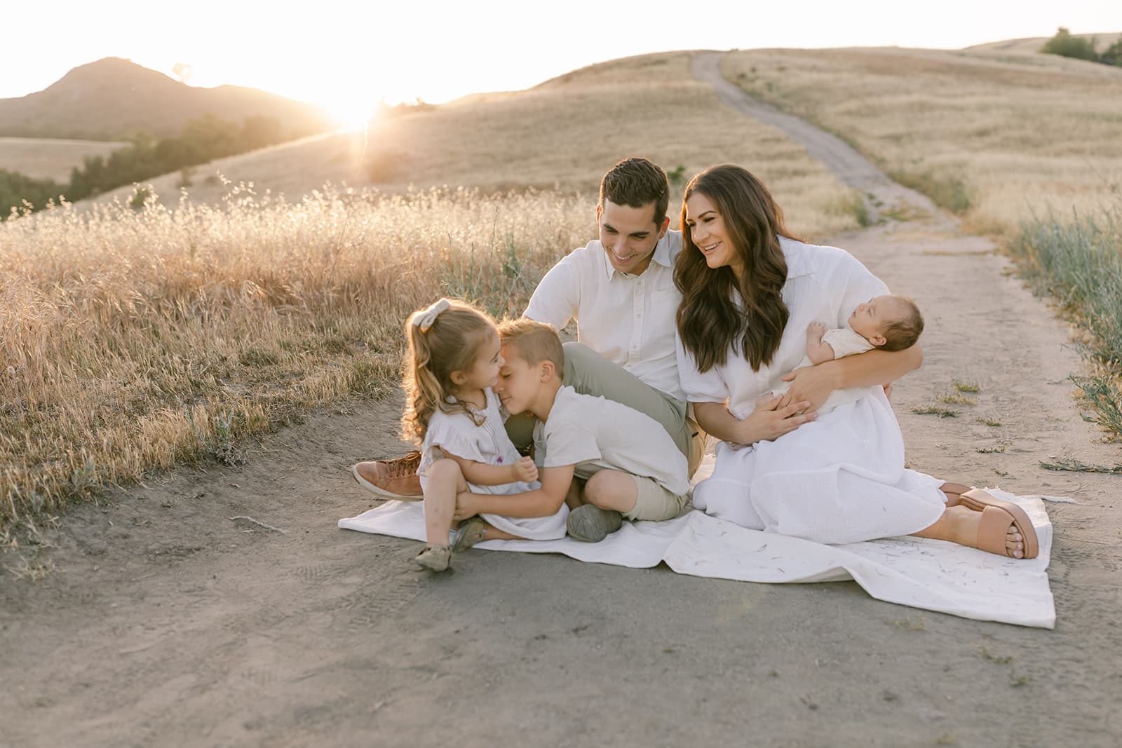 A mother and father sit on a blanket in a trail through rolling hills of grass playing with their three young children at sunset after meeting Newport Beach Nannies