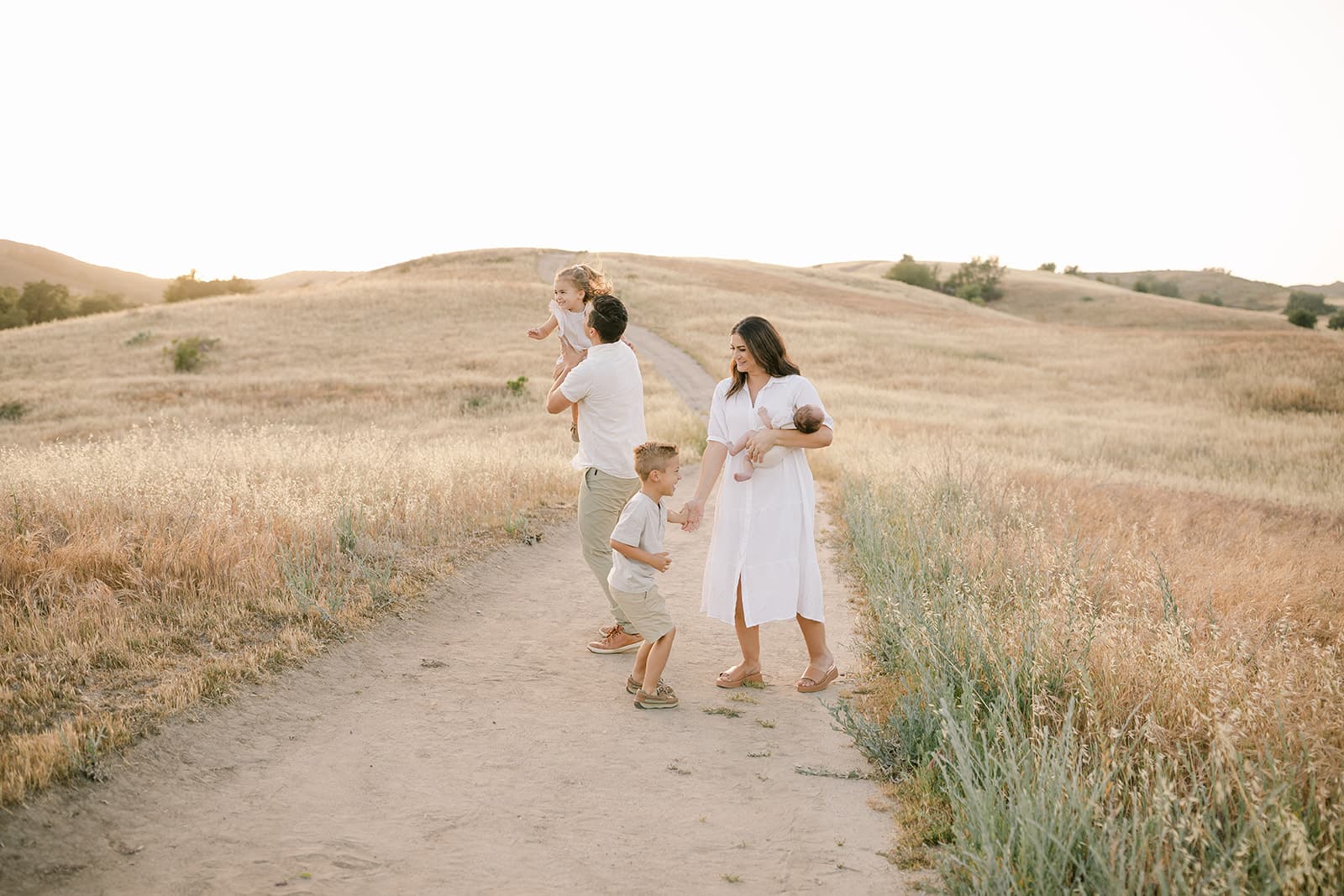 A mother and father in white dance and play with their newborn baby and two toddlers in a field of golden grass after meeting Newport Beach Nannies