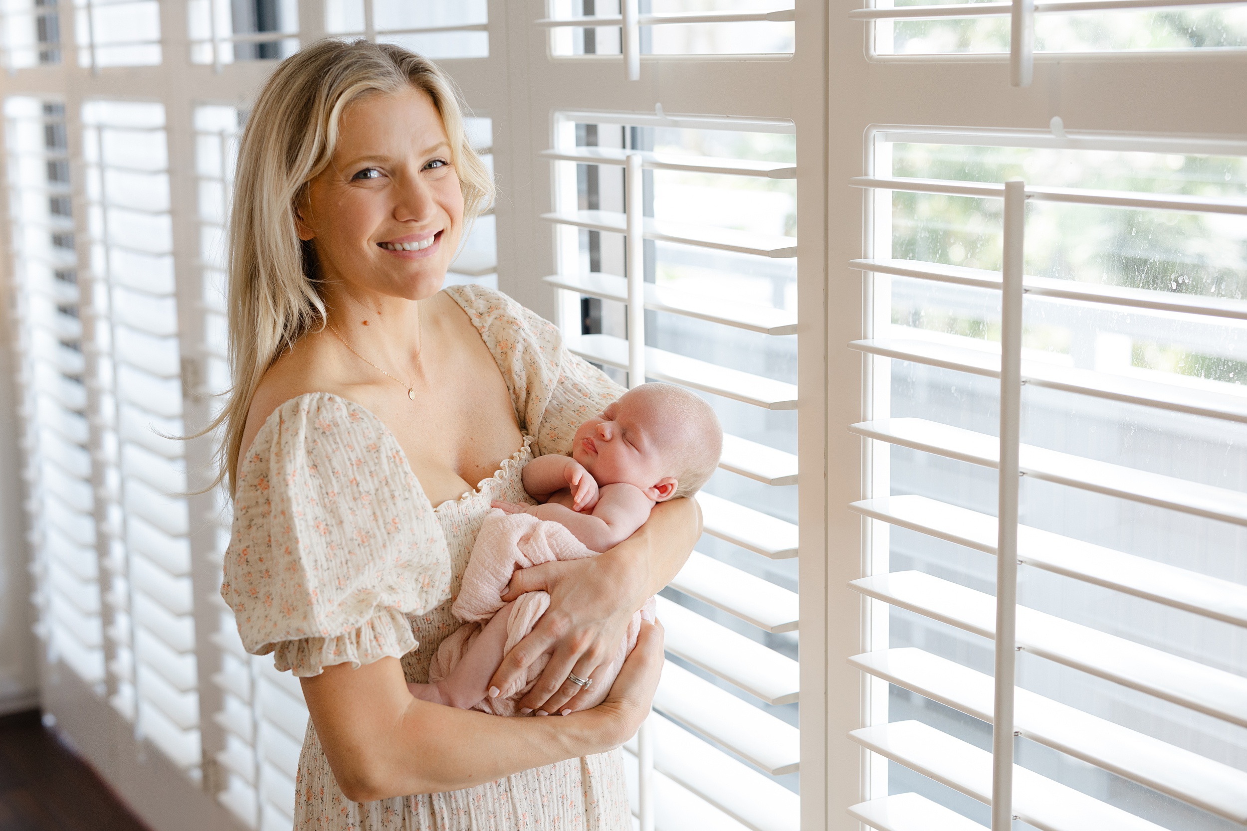 A happy new mother stands in a window in a floral print dress holding her sleeping newborn baby thanks to baby sleep consultant orange county