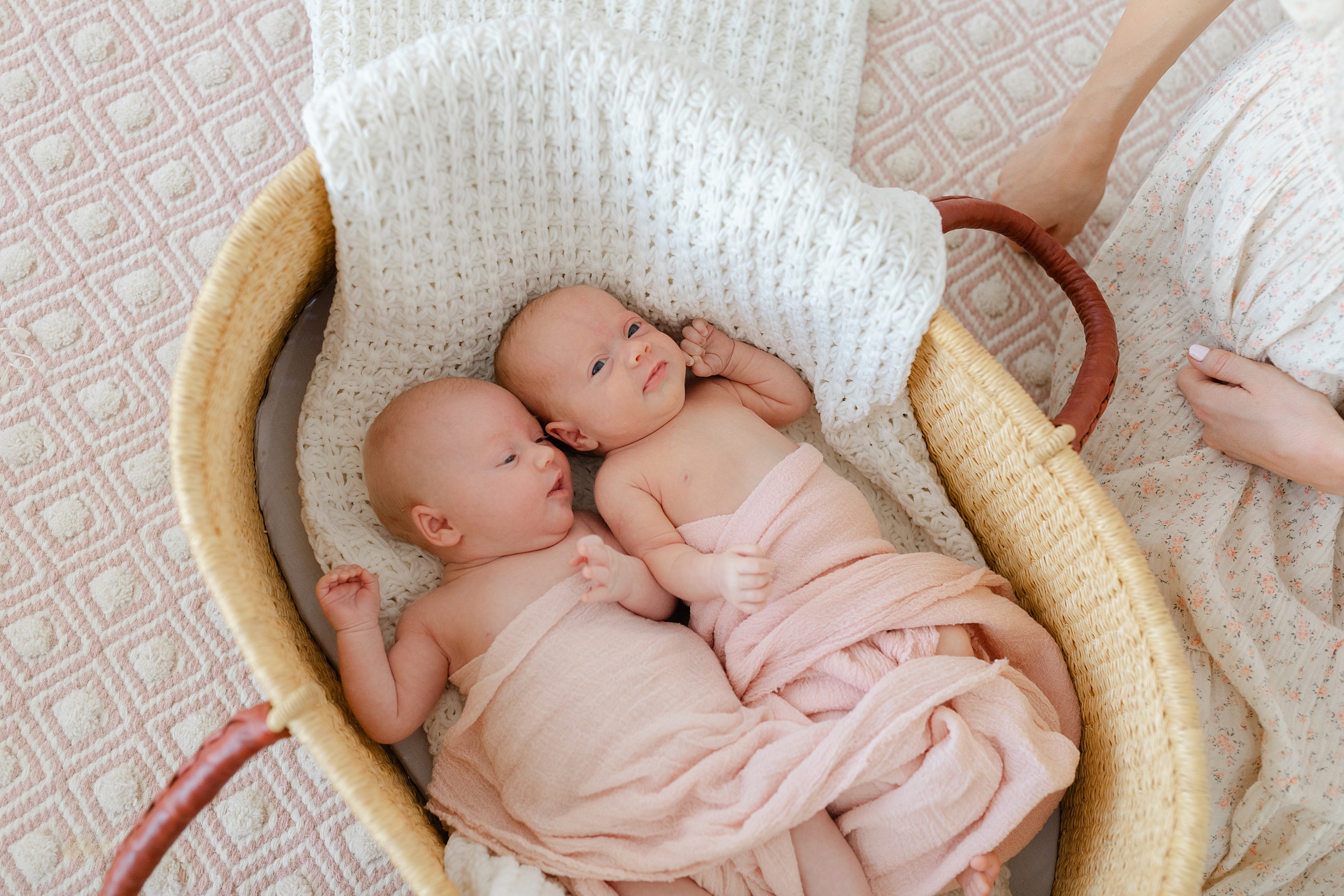Twin newborn babies lay awake in a woven basket with mom watching over them after meeting baby sleep consultant orange county