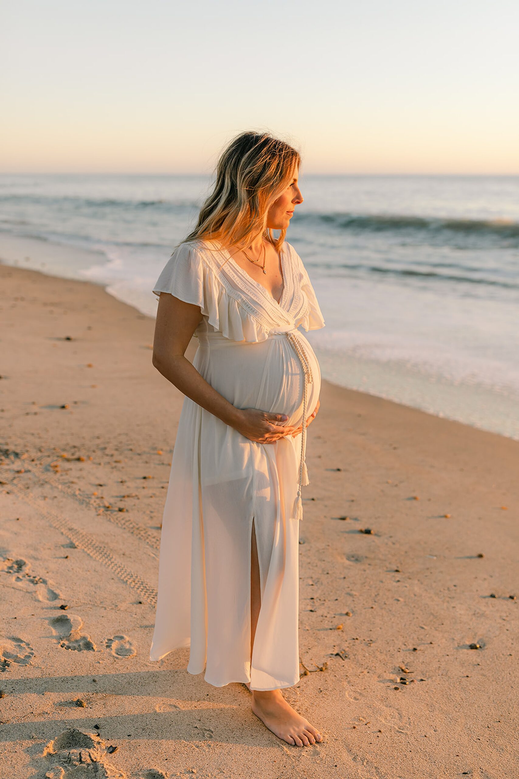 A mother to be in a white maternity gown walks down a beach at sunset holding her bump