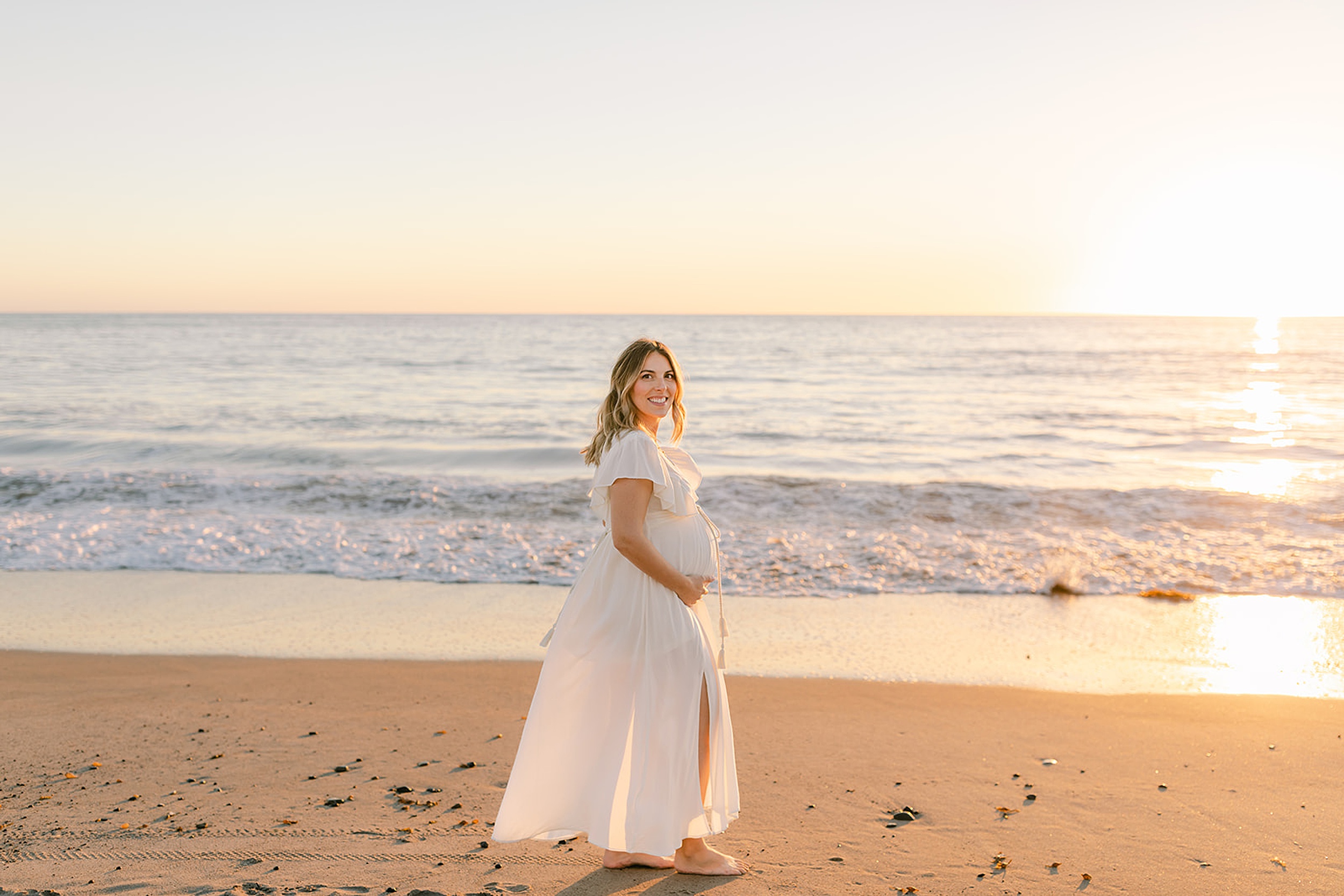 A mother to be in a white maternity gown stands on a beach at sunset holding her bump