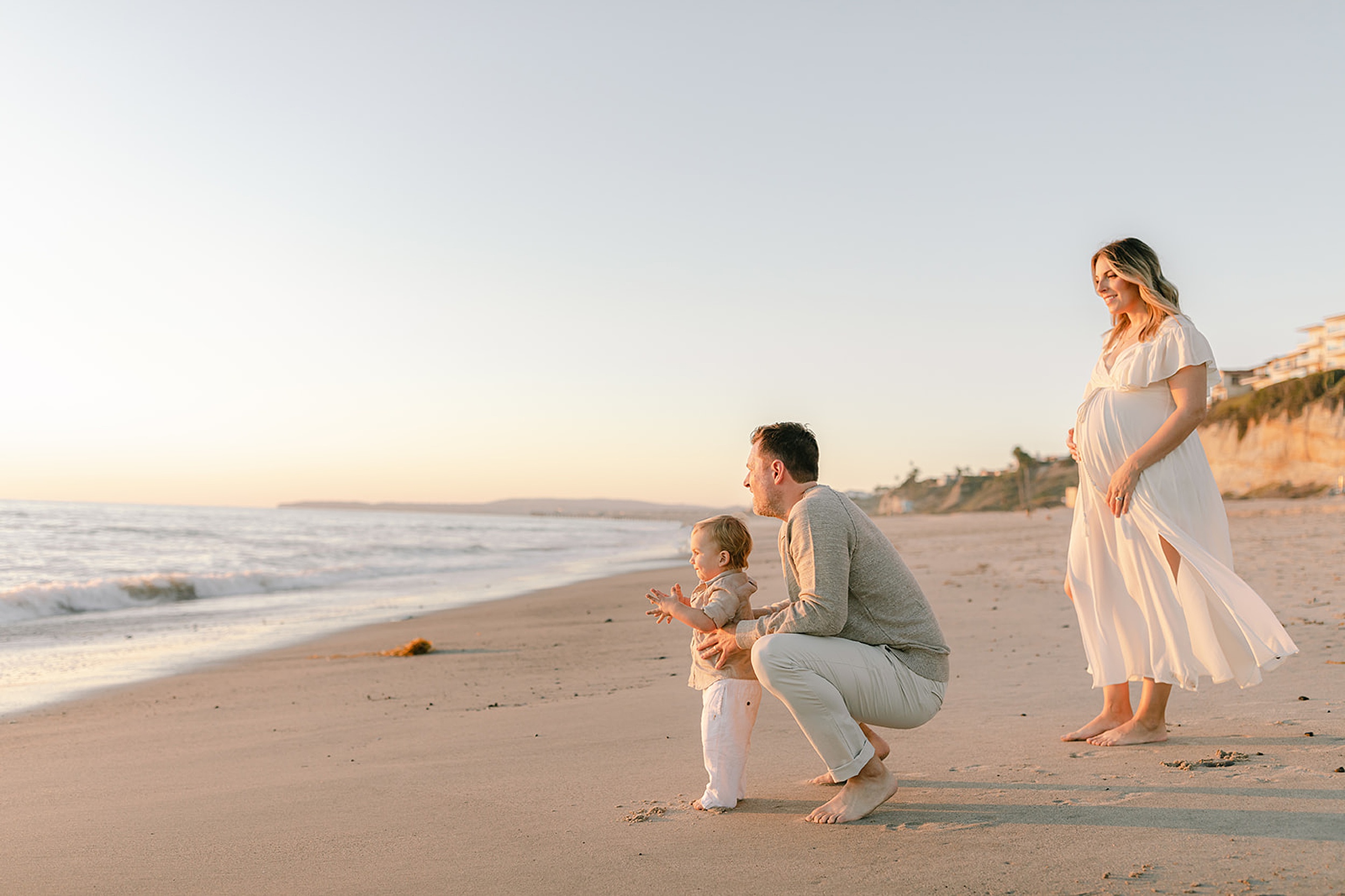 A pregnant mother stands on a beach watching her husband explore the sunset with their toddler son thanks to fertility clinics in orange county ca