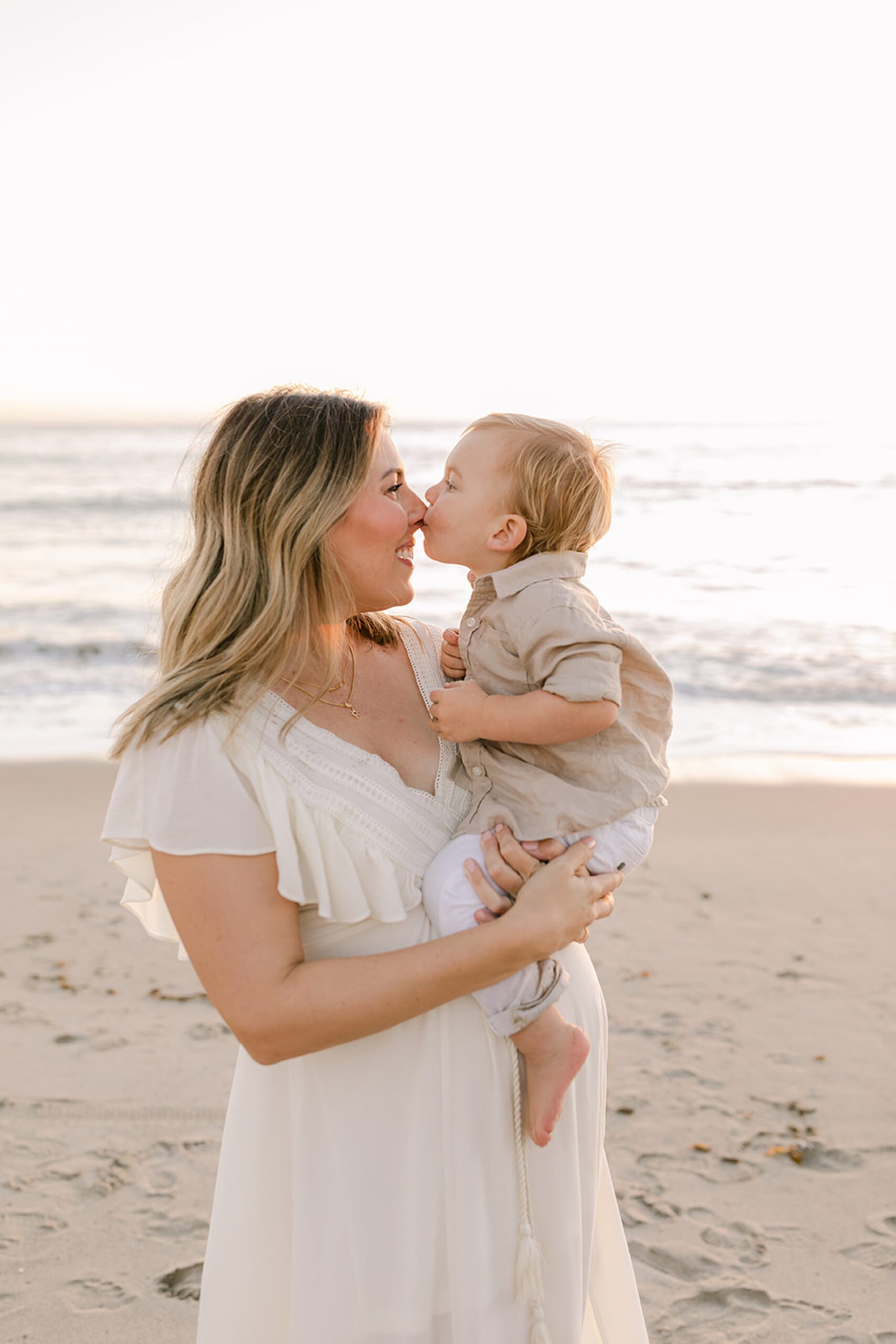 A mother stands on a beach in a white dress while her toddler son kisses her nose at sunset thanks to fertility clinics in orange county ca