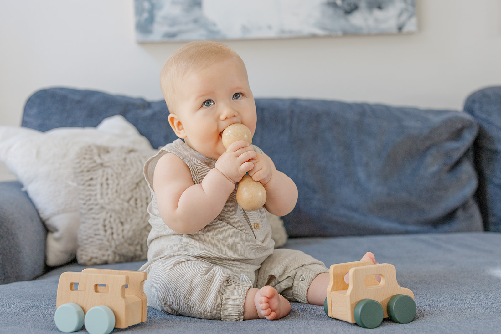 A toddler boy in a grey onesie chews on a wooden toy while sitting on a couch before meeting Irvine Pediatricians