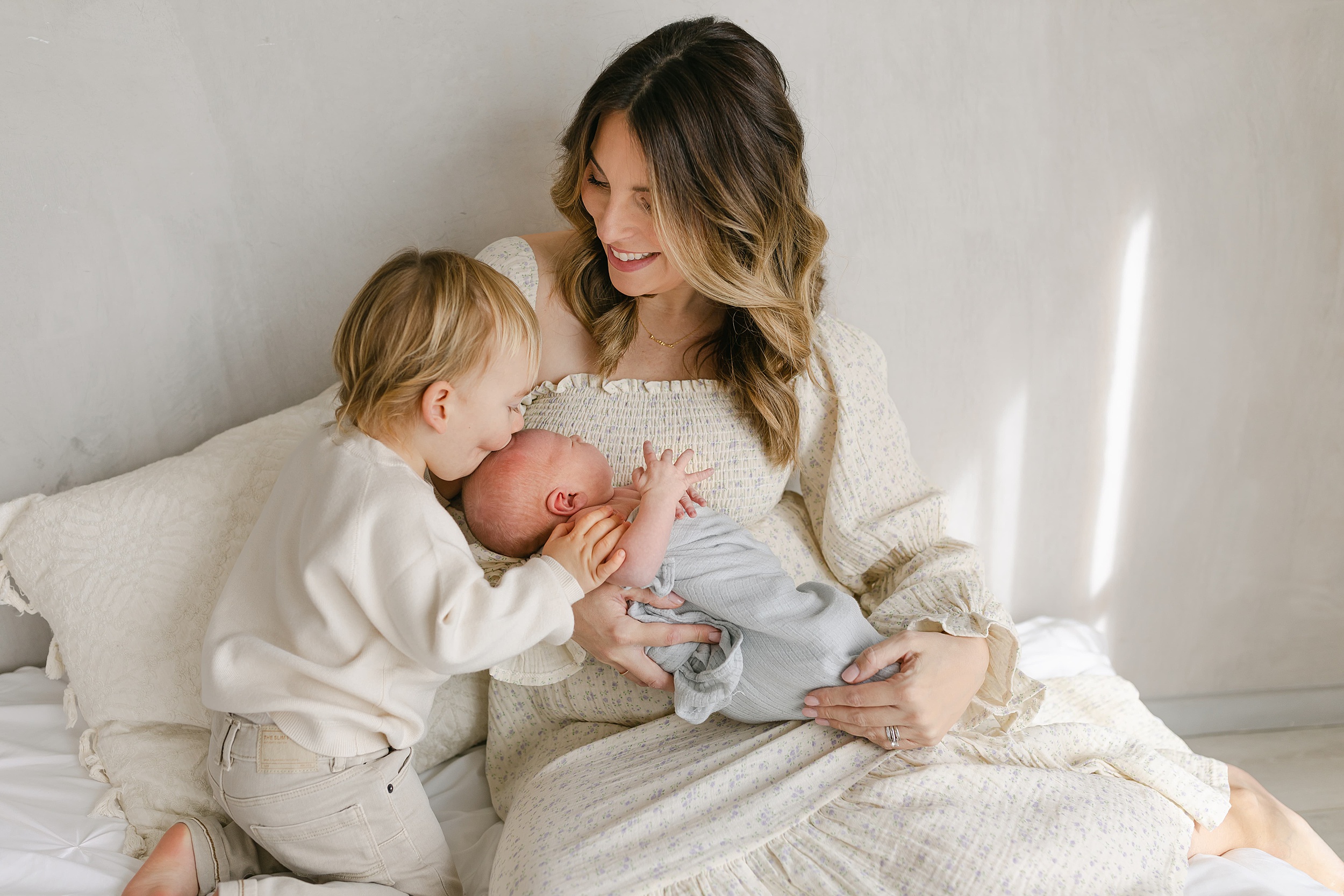 A young toddler kisses the head of their newborn sibling in mom's arms while sitting on a bed