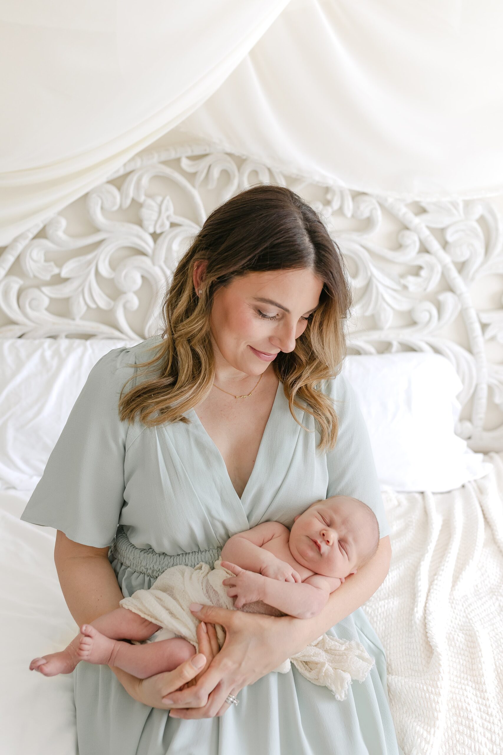 A happy new mother sits on a white bed smiling down at her sleeping newborn baby in her arms thanks to a lactation consultant orange county