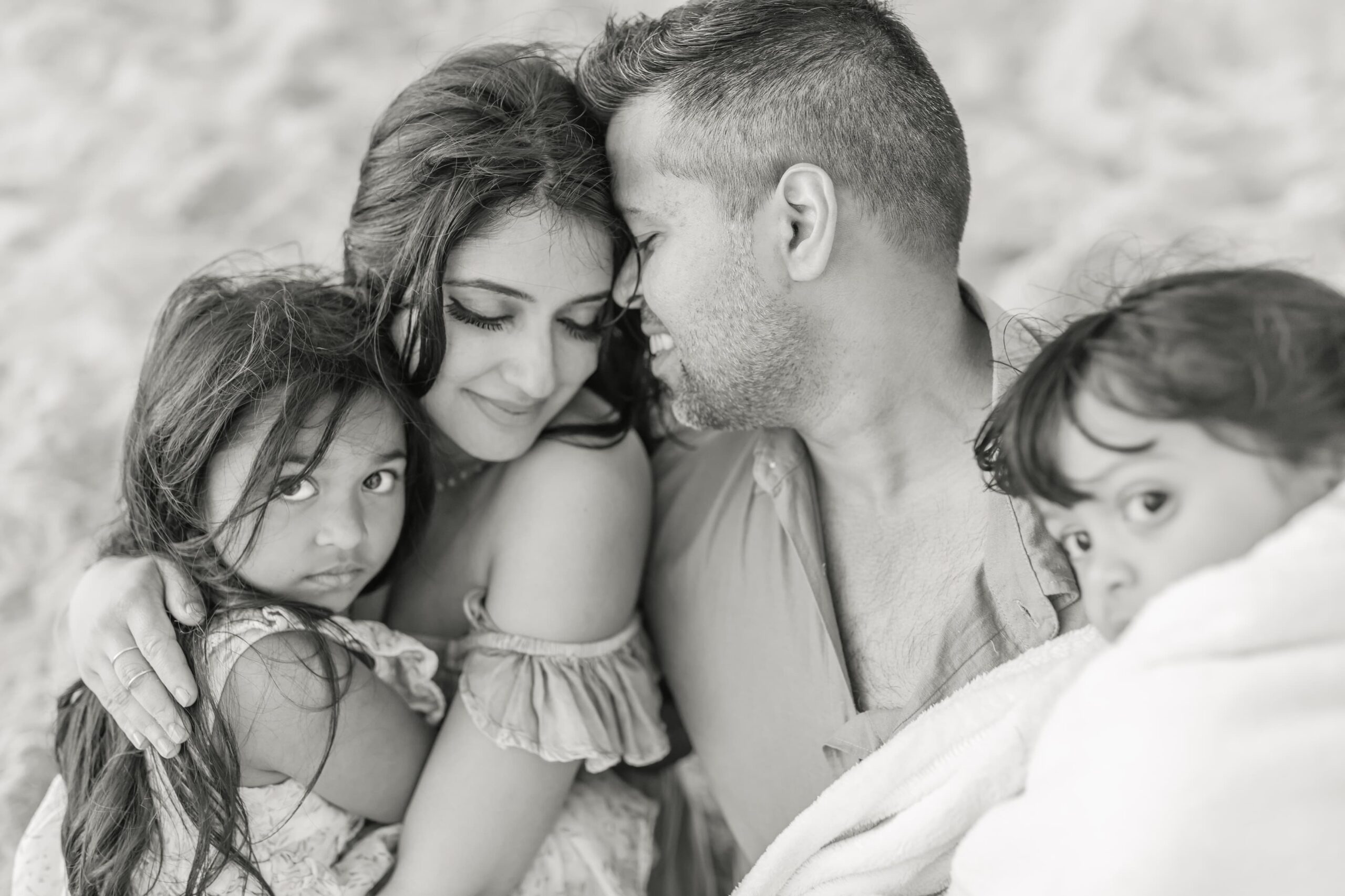 Happy parents snuggle on a beach with their toddler daughters