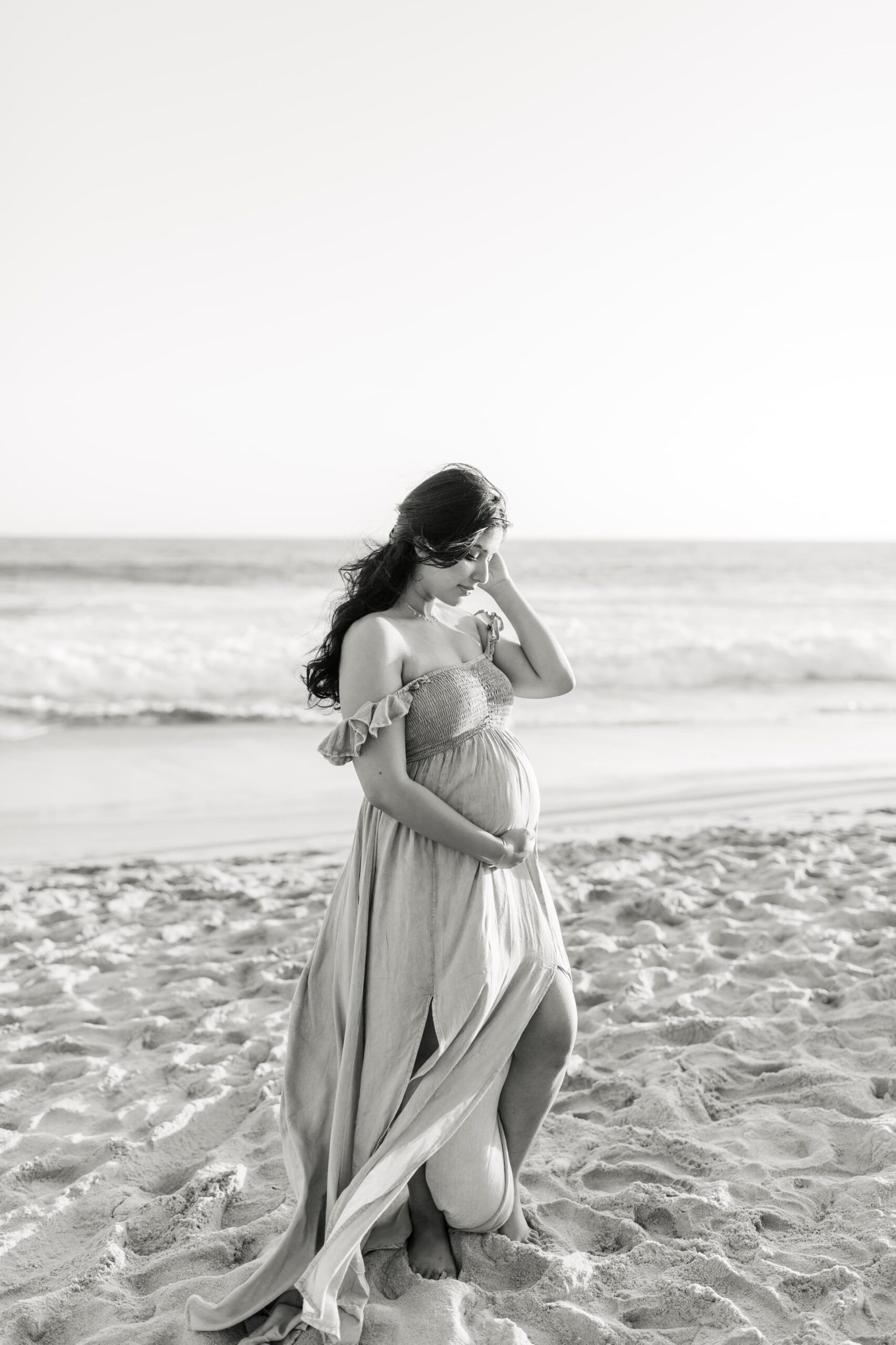 A mother to be in a flowing maternity dress stands on a beach in black and white before some lamaze classes orange county