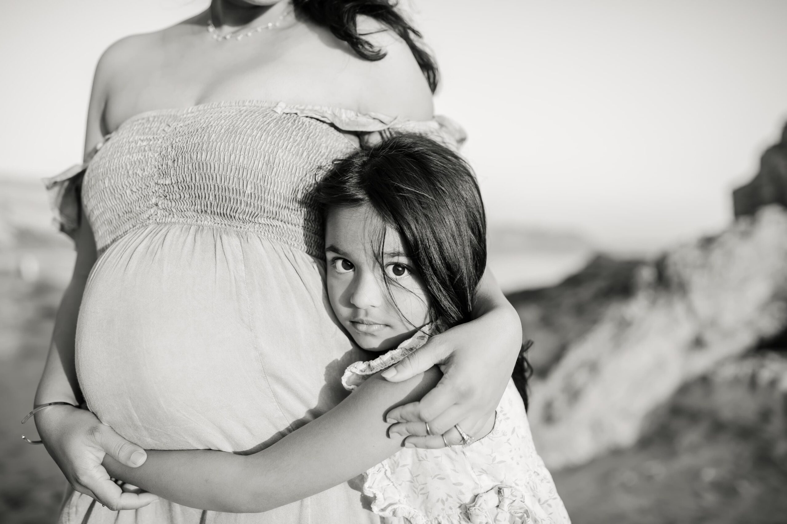 A young girl in a floral print dress hugs her pregnant mother while standing outside before some lamaze classes orange county