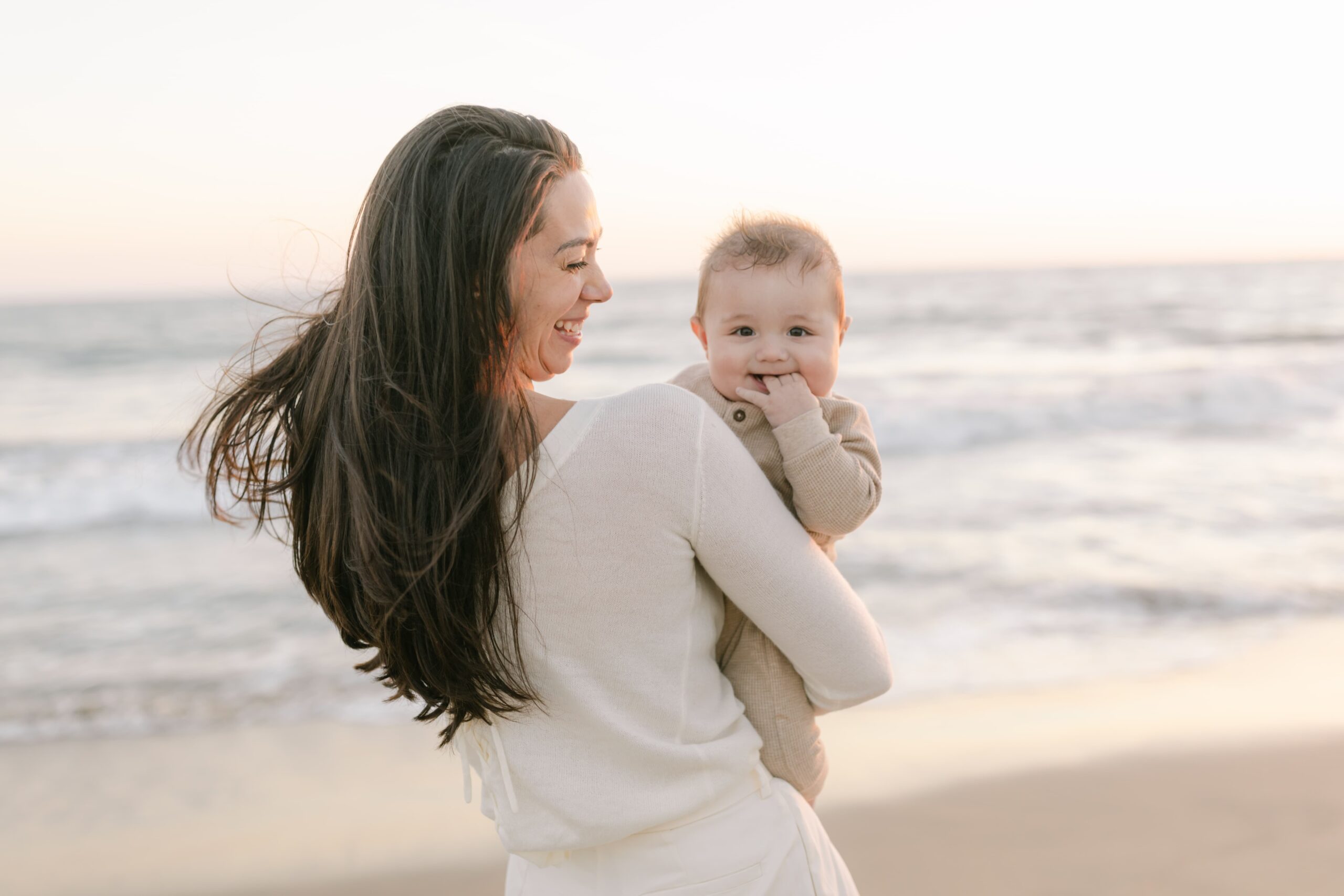 A happy mother smiles at her infant son sitting in her arms while standing on a beach at sunset