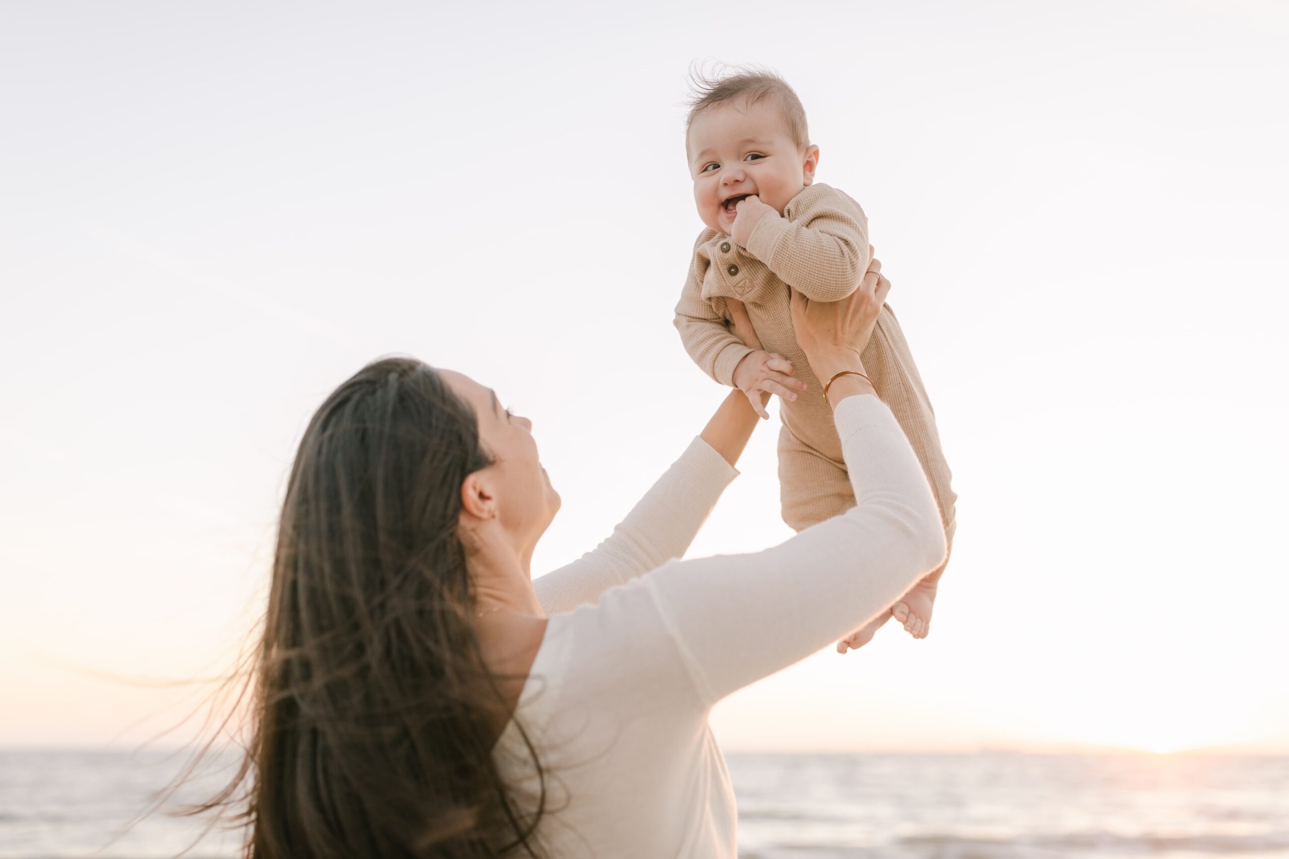 A happy mother lifts her giggling infant son in a tan onesie on a beach at sunset after some Mommy and Me Classes Orange County