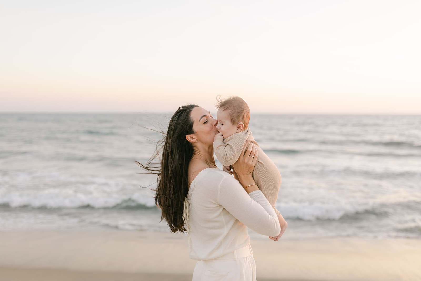 A mother in a white outfit stands on a windy beach kissing her infant son in a brown onesie after some Mommy and Me Classes Orange County