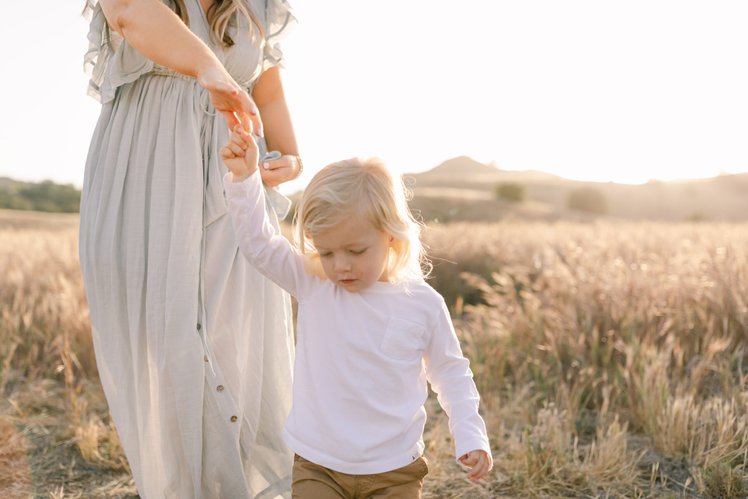 A toddler boy leads his mom by the hand through a park trail of tall golden grass