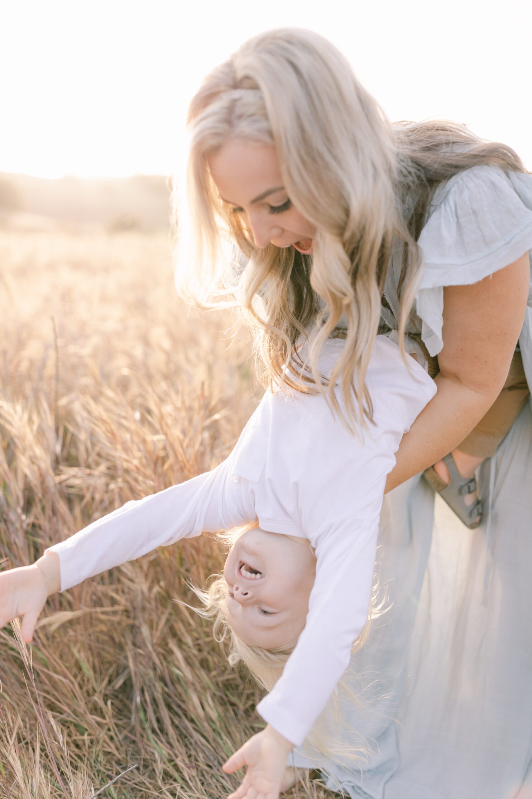 A mother in a blue dress tips her son upside down while playing in a field of tall golden grass after visiting Newport Beach Montessori