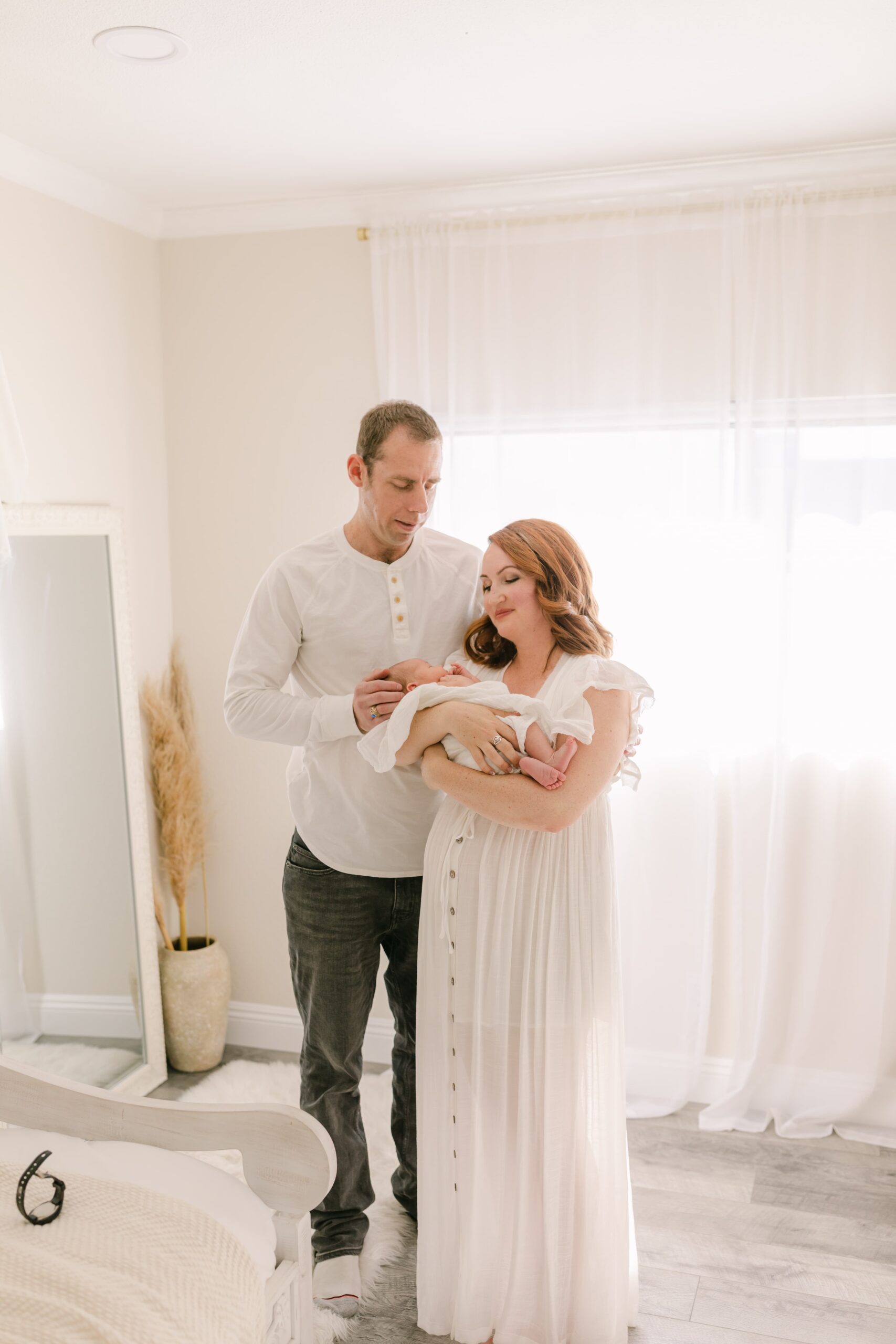 Happy new parents stand in a studio in white while cradling their sleeping newborn baby after finding a newport beach pediatrician