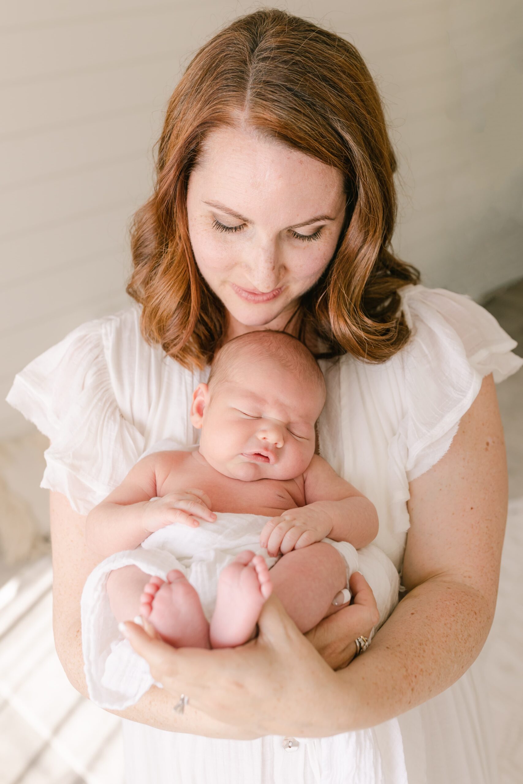 A new mother in a white dress cradles her sleeping newborn baby against her chest while standing in a studio before meeting a newport beach pediatrician