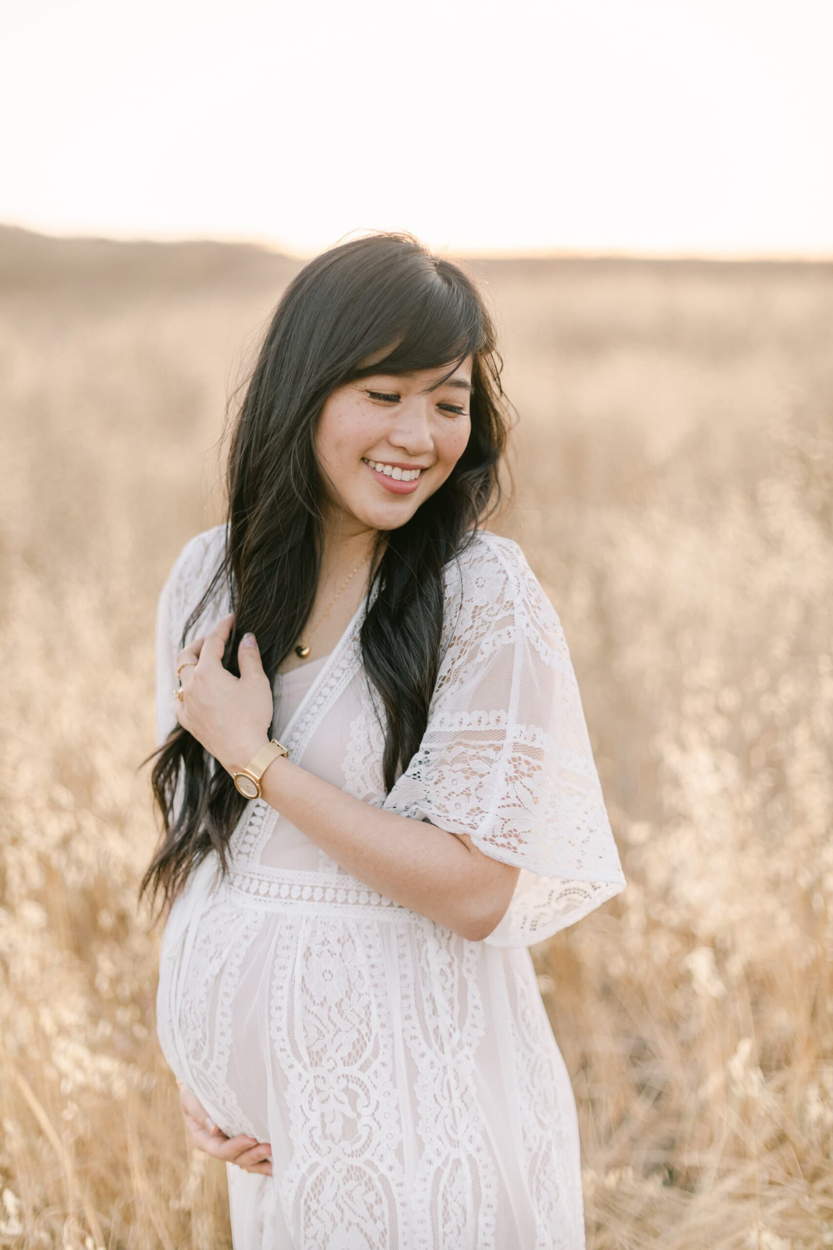 A mom to be in a white lace maternity gown smiles over her shoulder while standing in a field of golden grain