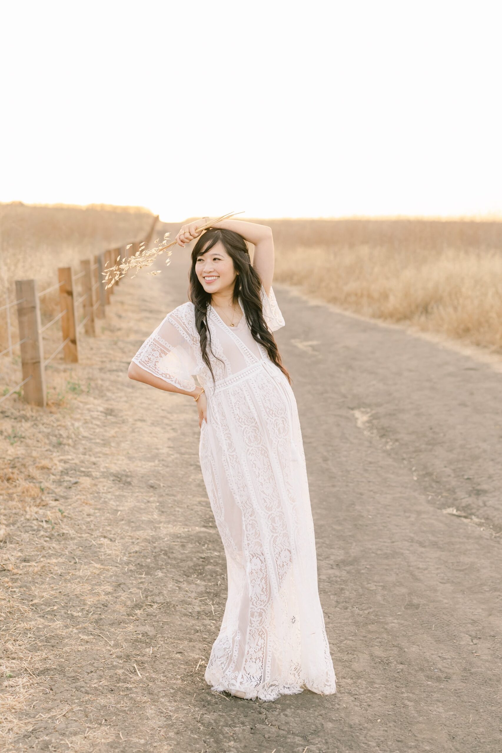 A mother to be in a white lace dress stands in a rural road through a field of golden grass at sunset smiling and holding grain