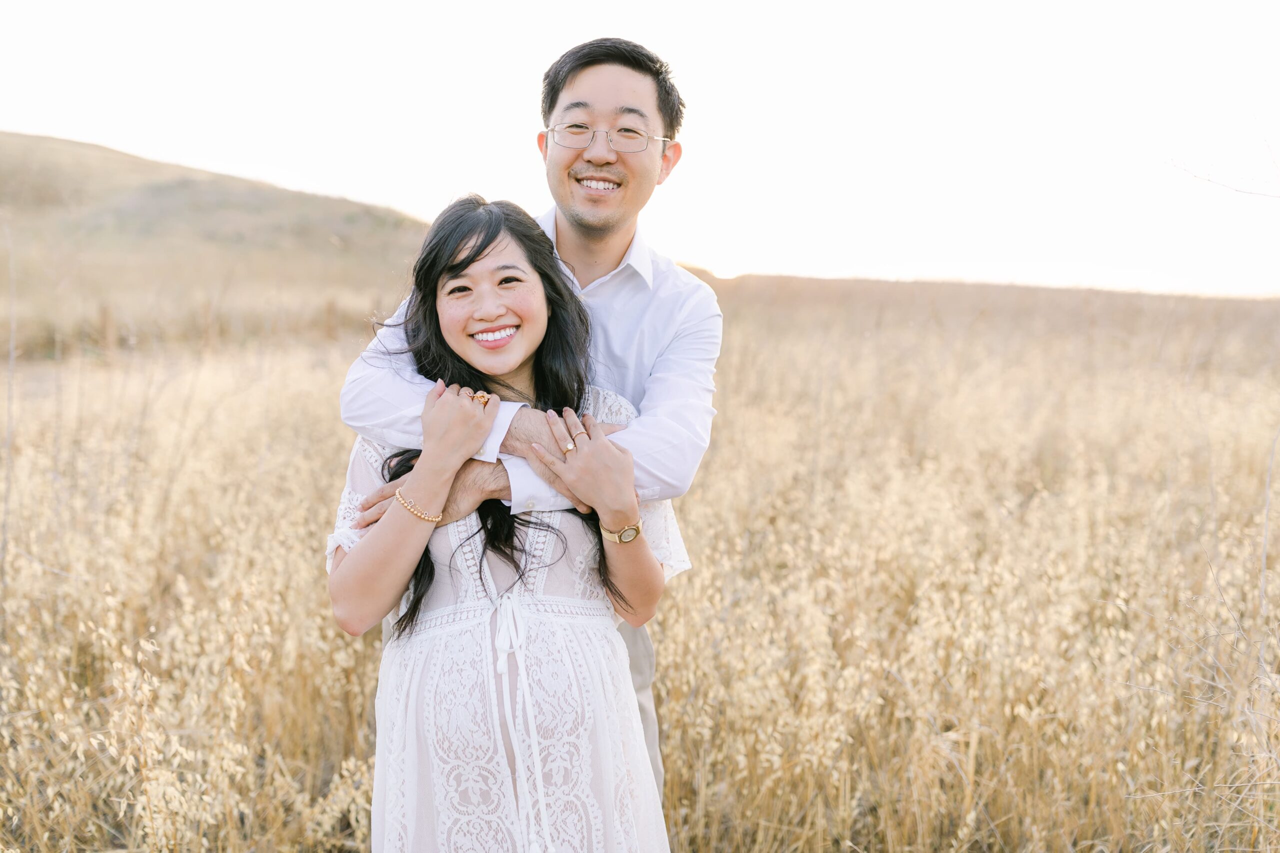 A husband hugs his pregnant wife from behind while standing in a field at sunset after finding an obgyn orange county