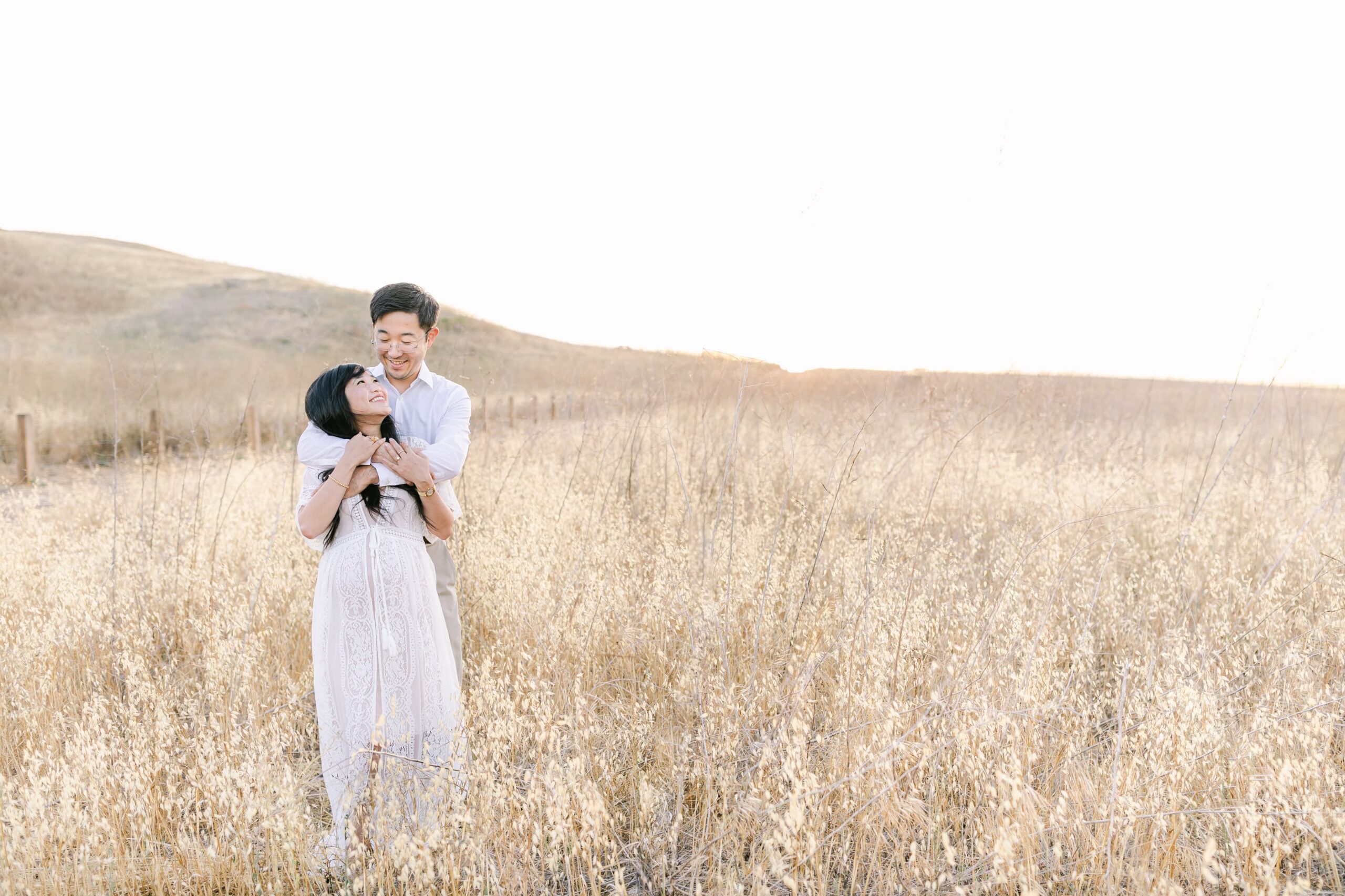 Happy expecting parents share a hug while standing in a field of tall golden grass at sunset after visiting obgyn orange county