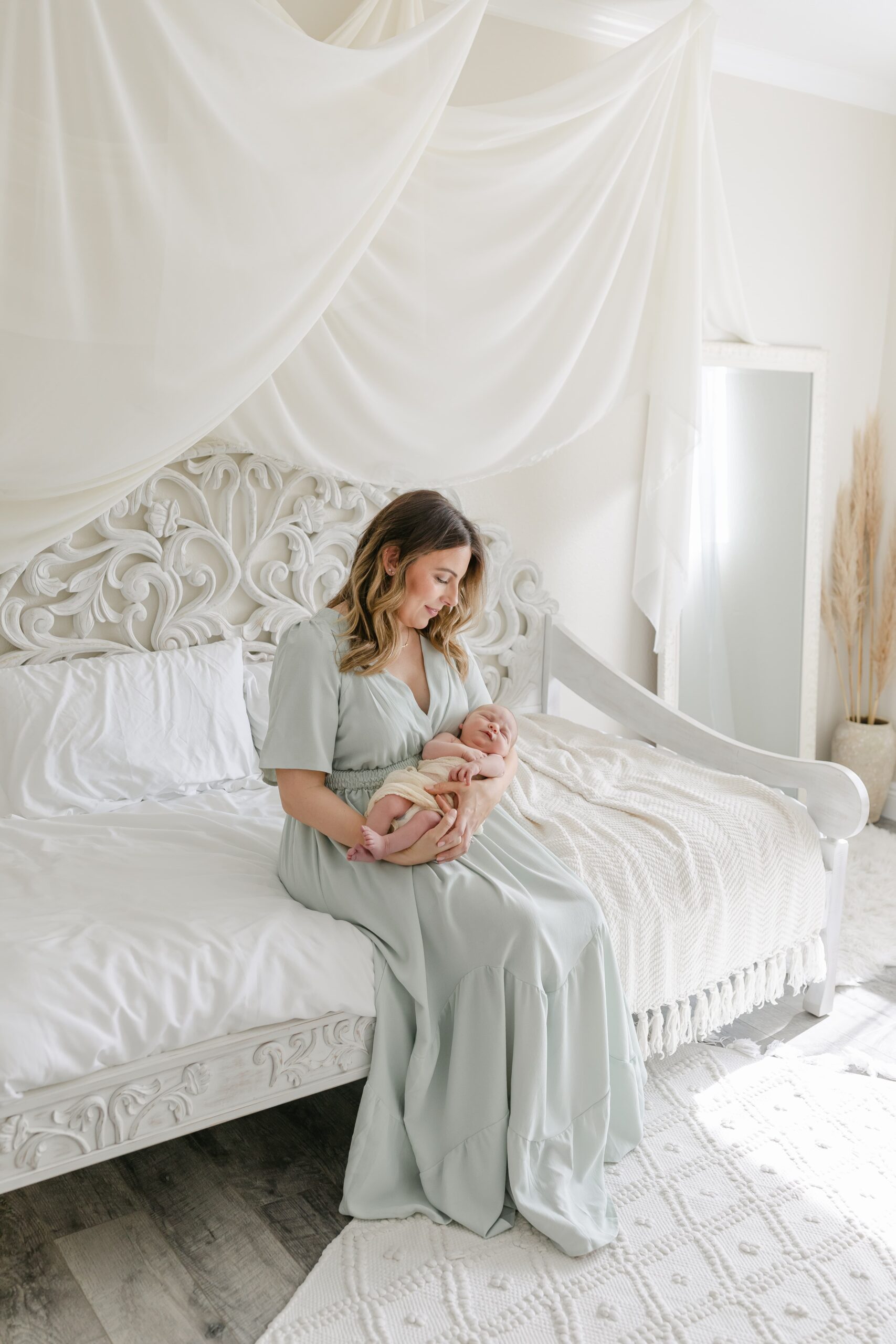 A new mother in a green dress sits on the edge of an ornate bed in a studio cradling her sleeping newborn baby