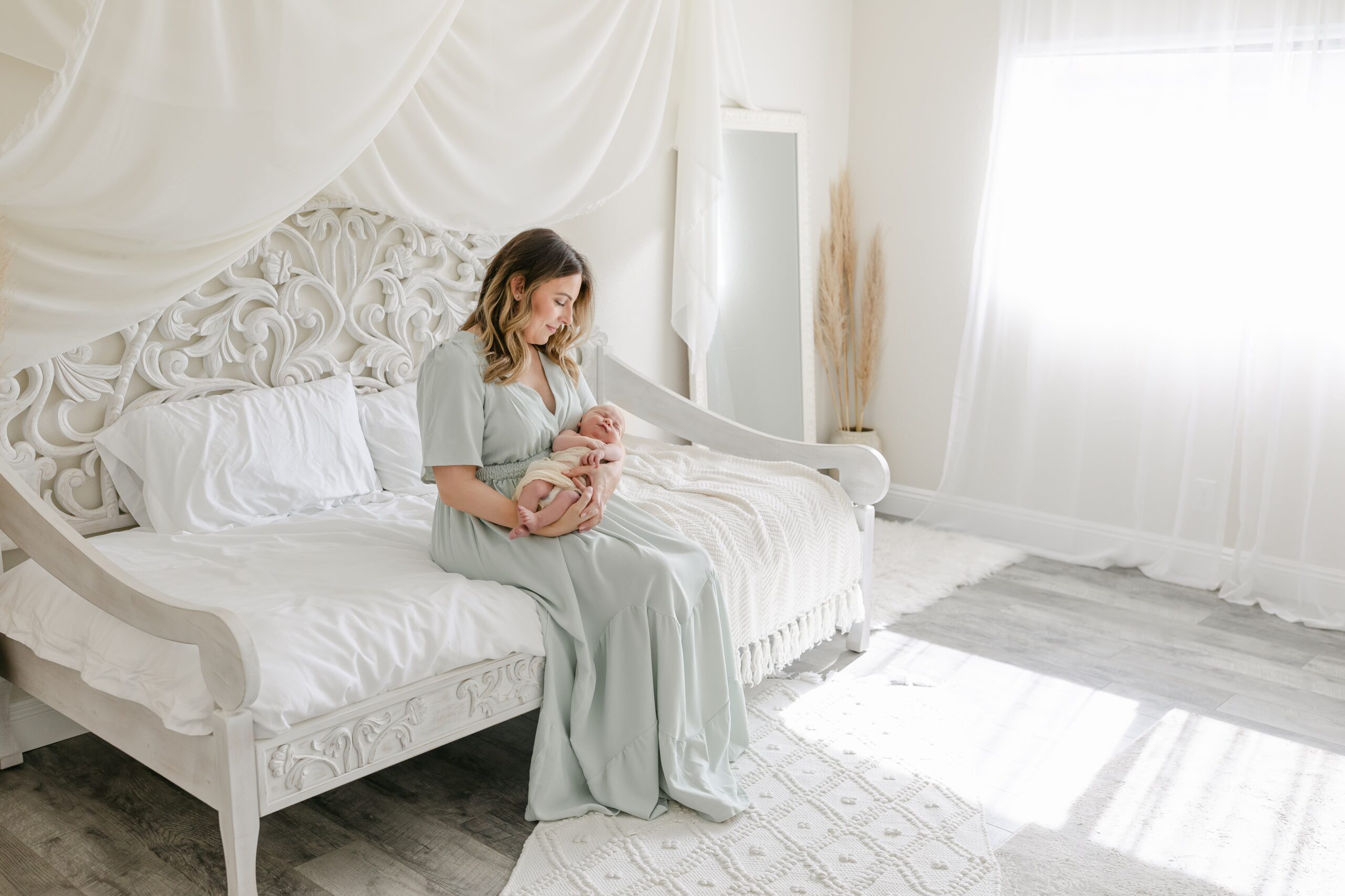 A happy new mother in a green dress sits on a day bed in a studio holding her sleeping newborn baby before visiting orange county baby boutiques