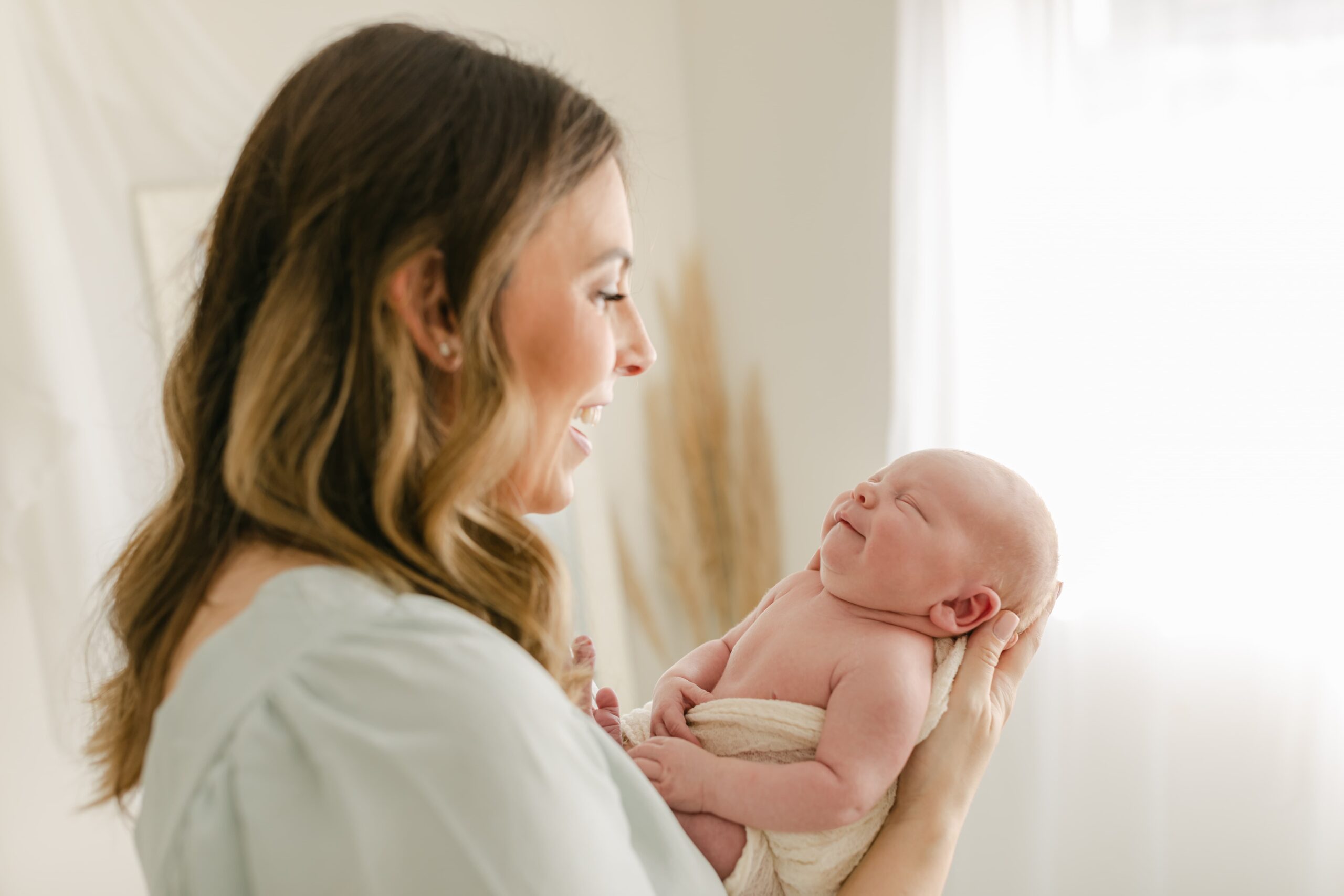 A happy mother laughs while standing in a studio holding her smiling sleeping newborn baby before visiting orange county baby boutiques