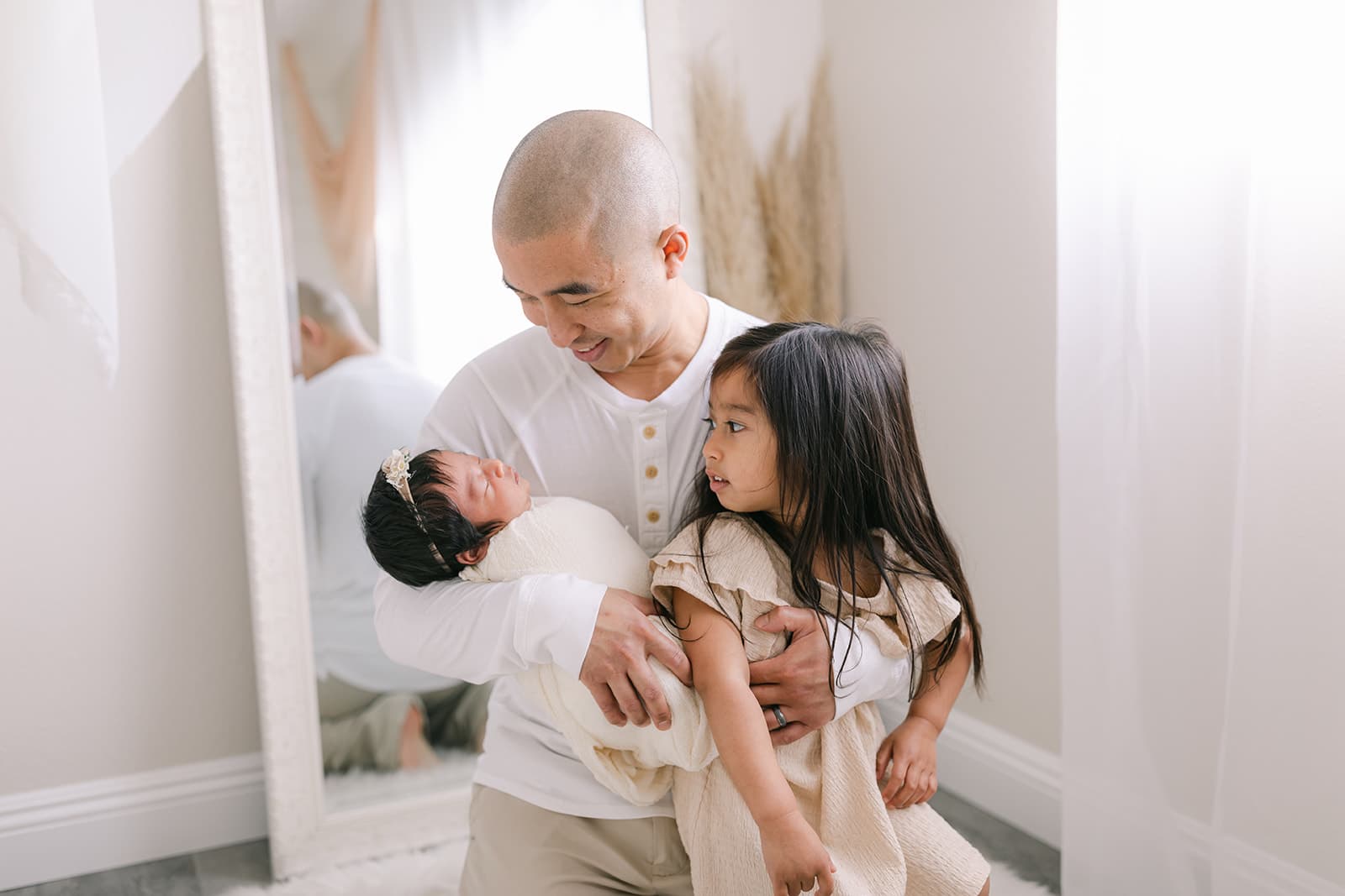 A happy father holds his sleeping newborn baby and toddler daughter in a white room in front of a mirror