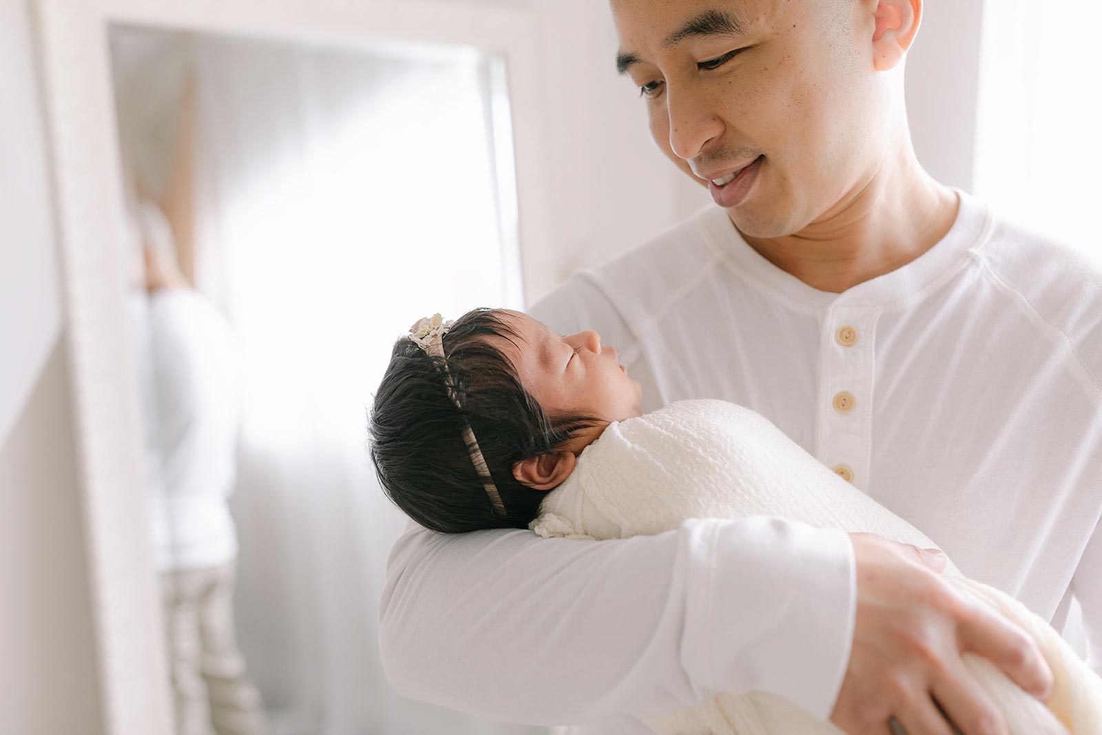 A happy father in a white henley smiles down at his sleeping newborn baby daughter in a white swaddle after using orange county birth center