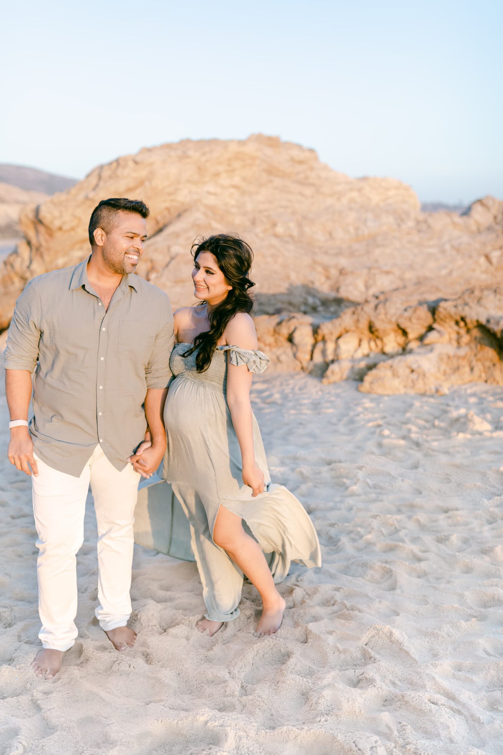 Happy expecting parents walk on a rocky beach at sunset holding hands in the wind after meeting orange county doulas