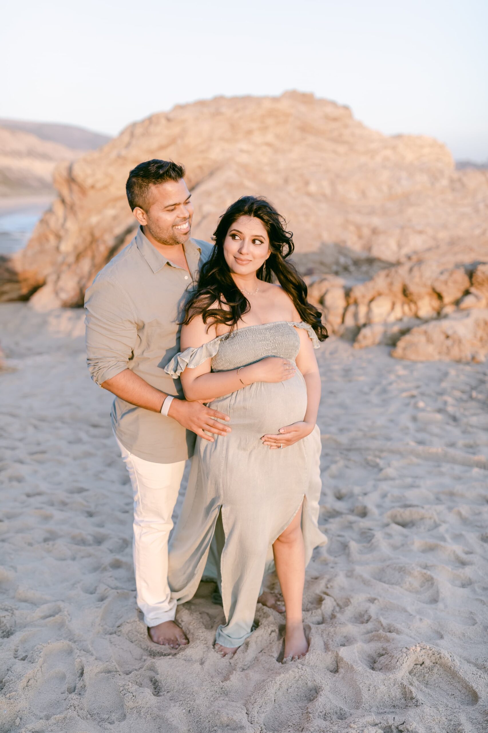 Happy expecting parents stand on a beach cuddling while holding the bump at sunset after meeting orange county doulas