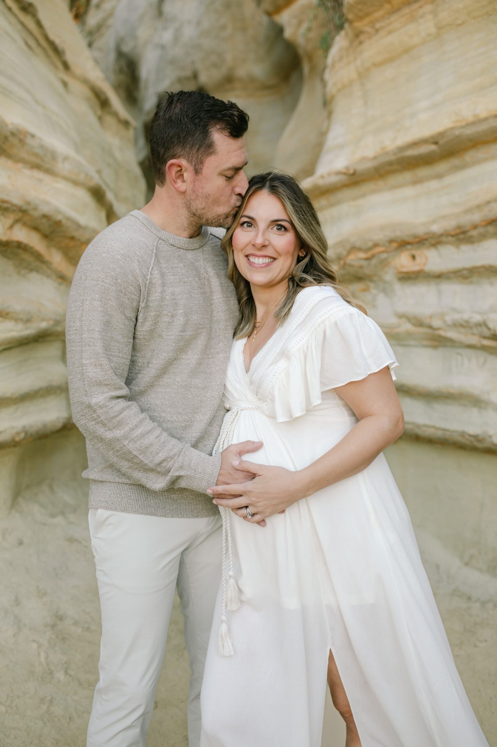 A father to be kisses his pregnant while while holding the bump and standing on a beach