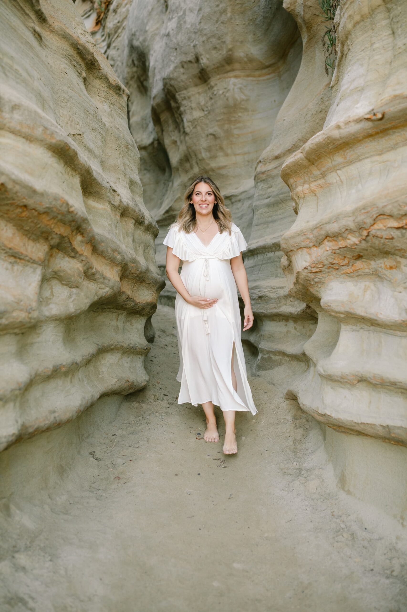 A mom to be walks down a gorge to the beach while smiling and holding her bump after meeting orange county midwives