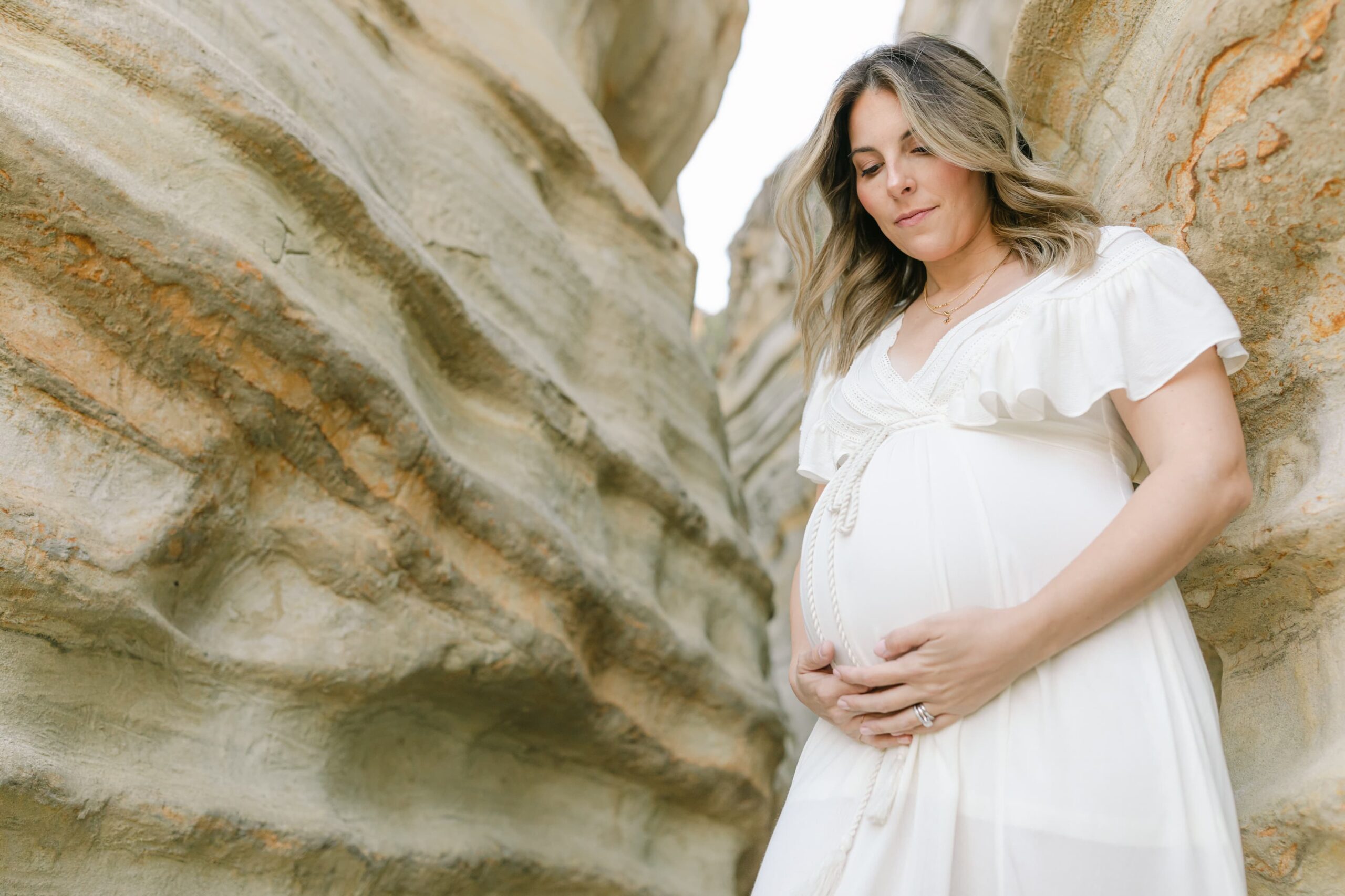 A mom to be in a white dress leans against the wall of a gorge holding the bump after meeting orange county midwives