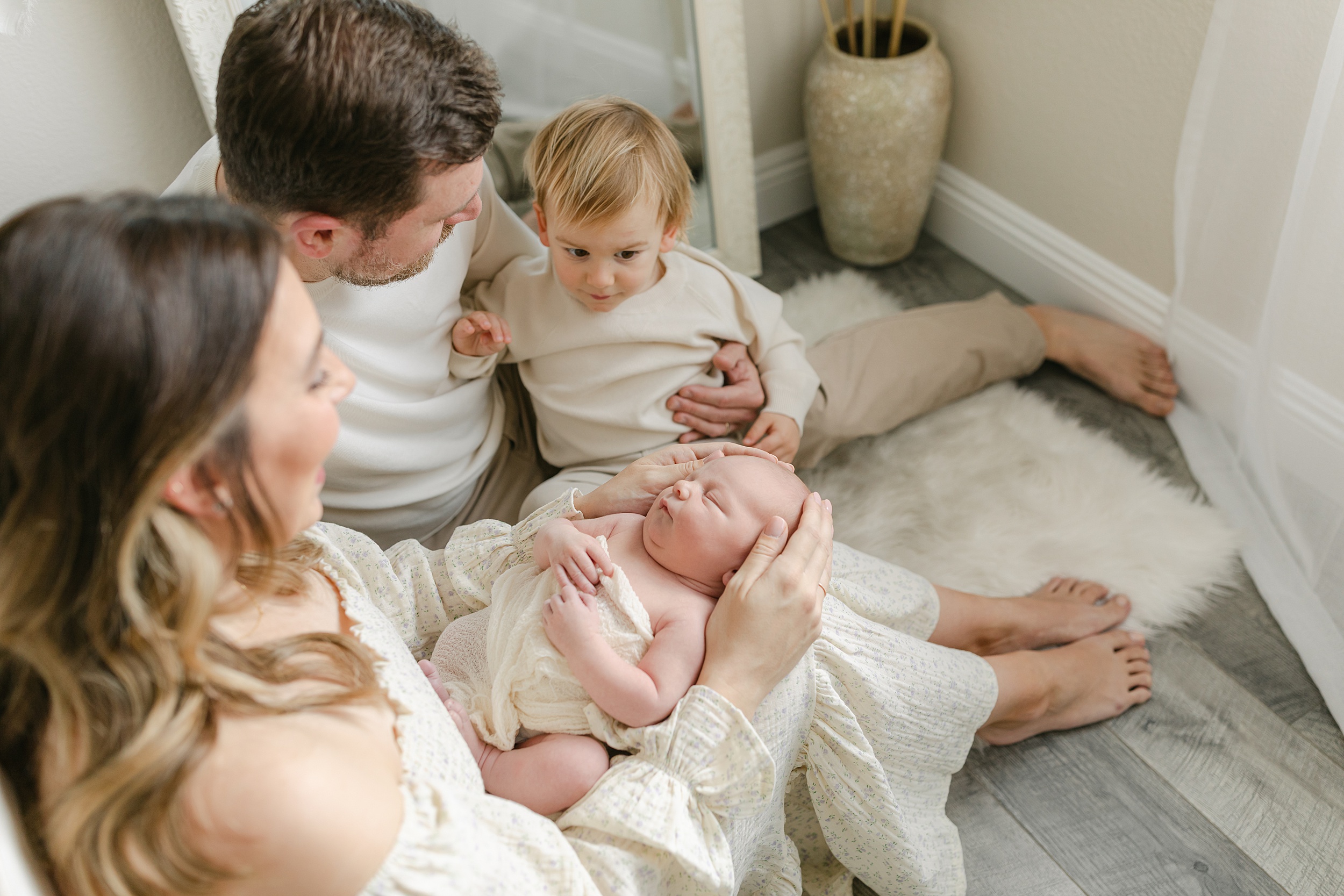 A mother and father play with their toddler son on the floor by a bed with their newborn sleeping in mom's lap